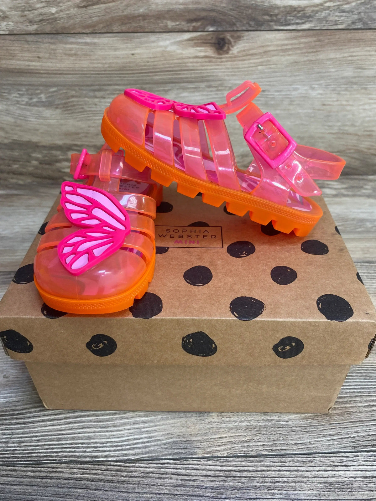 Side and front view of Sophia Webster jelly sandals in pink-orange with pink butterfly charm and buckle on polka-dot shoebox