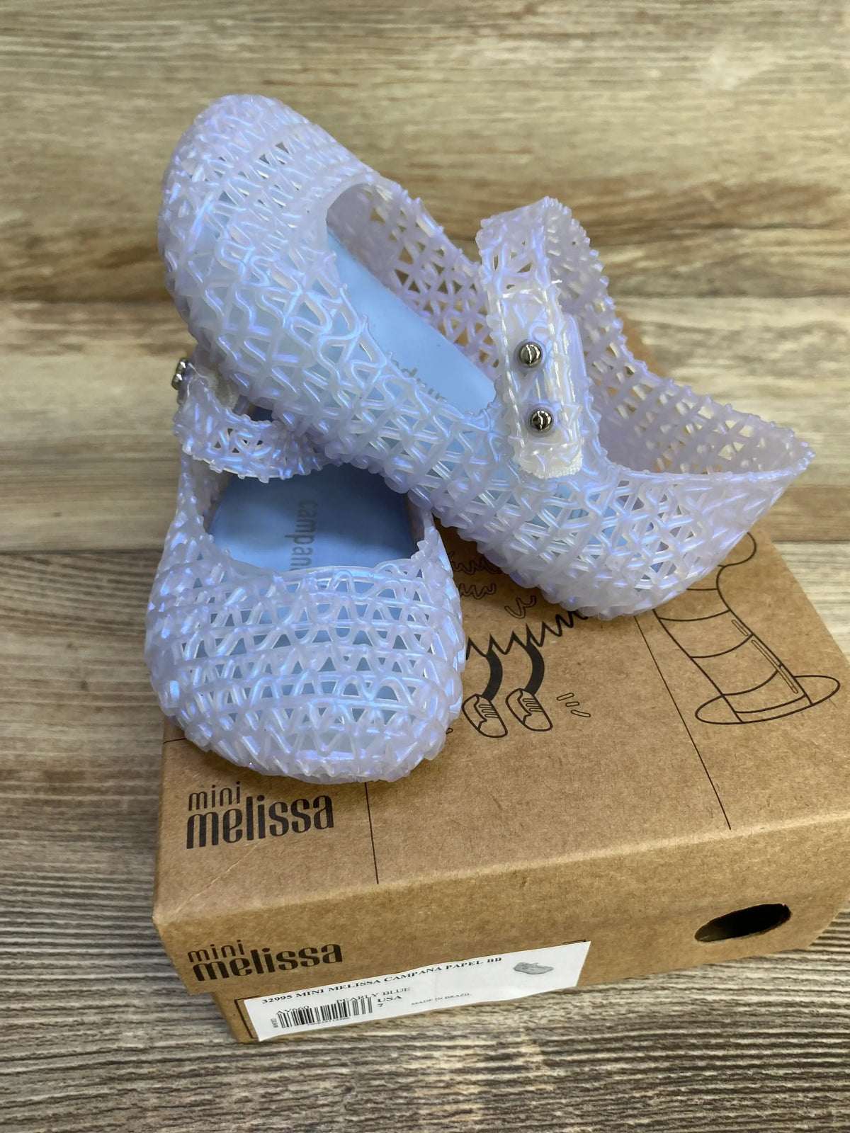 Three-quarter view of new Mini Melissa Campana Papel jelly flats in pearly blue sz 7c on top of original Mini Melissa box on wood plank floor