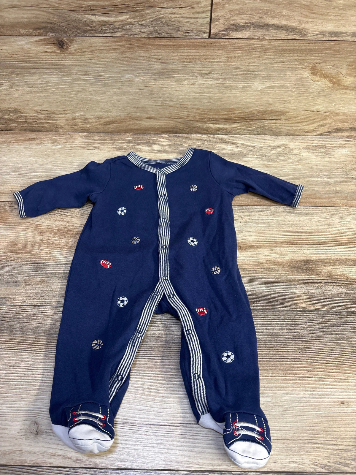 Top-down view of Little Me navy footed sleeper with embroidered soccer, basketball, and football motifs, striped trim, and sneaker-style feet on wooden surface