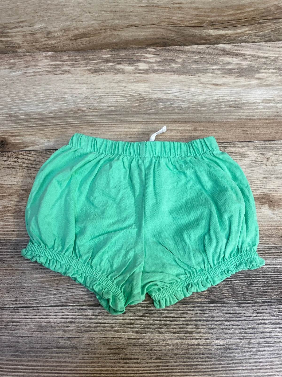 Top view of green baby shorts (3-6M) with elastic waistband, white drawstring, and ruffled leg openings on wood background