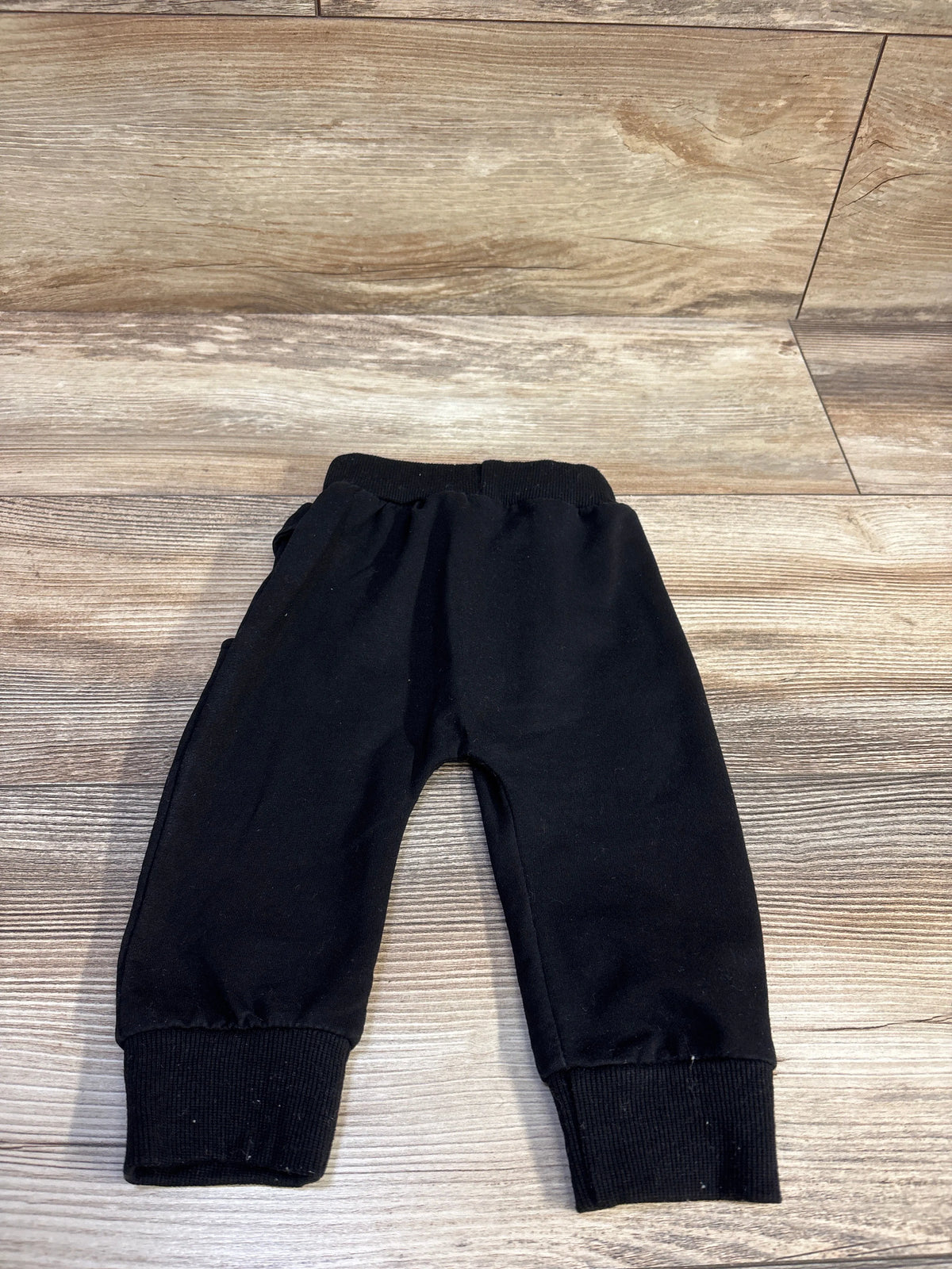 Flat lay of black drawstring harem pants size 3-6 months showing elastic waistband and ribbed ankle cuffs on wood background