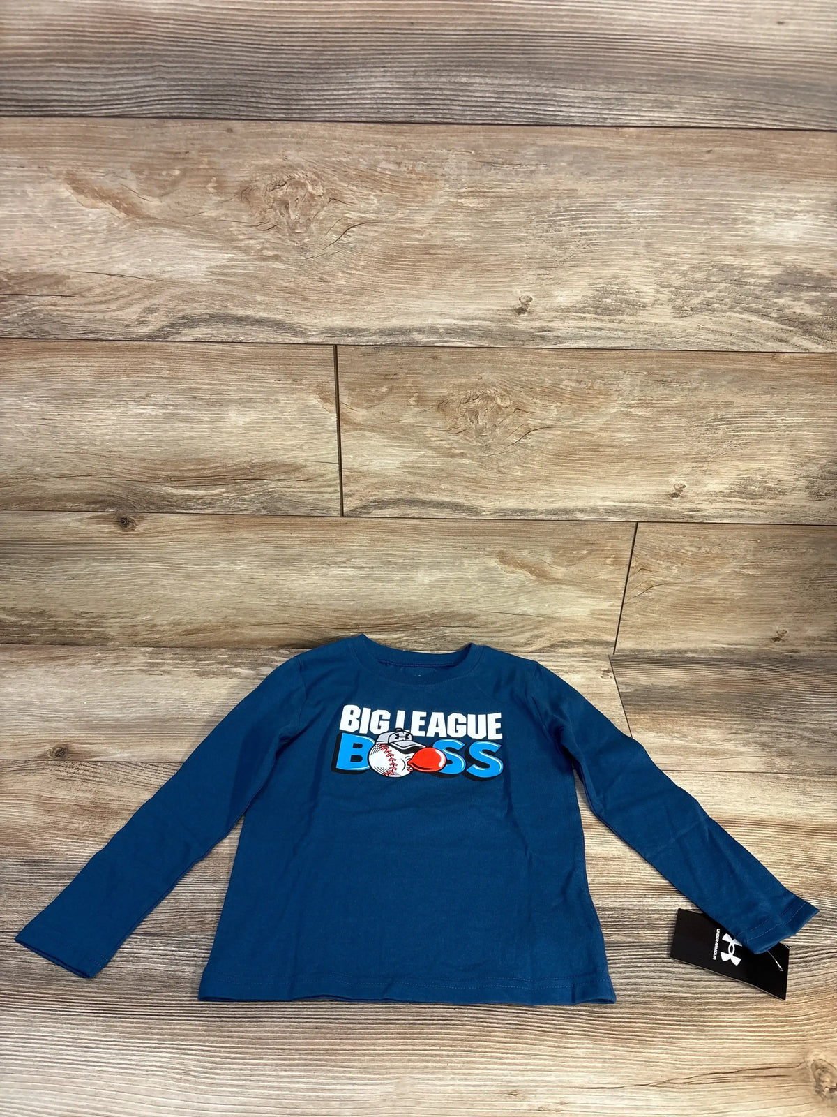 Flat lay of blue Under Armour 4T long-sleeve shirt showing BIG LEAGUE BOSS text and baseball graphic on wood background