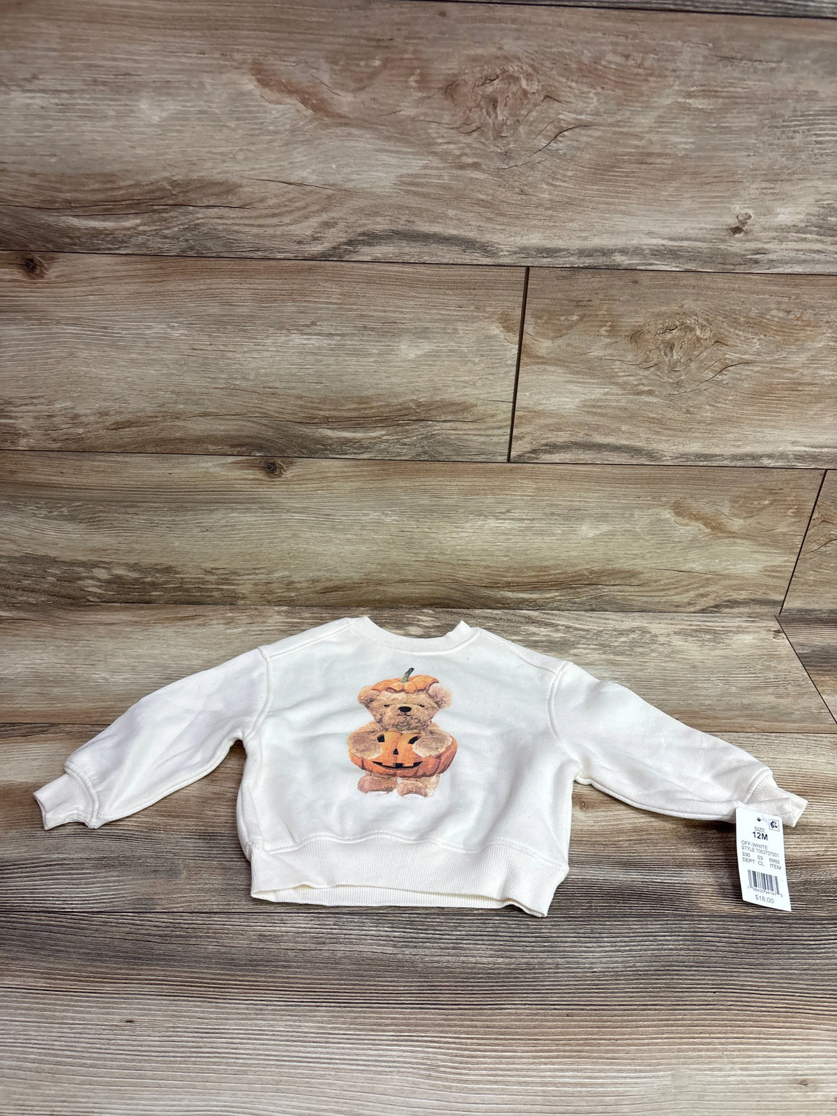 Front view of the ivory Grayson Mini Pumpkin Bear sweatshirt with long sleeves and pumpkin bear graphic on a wood floor