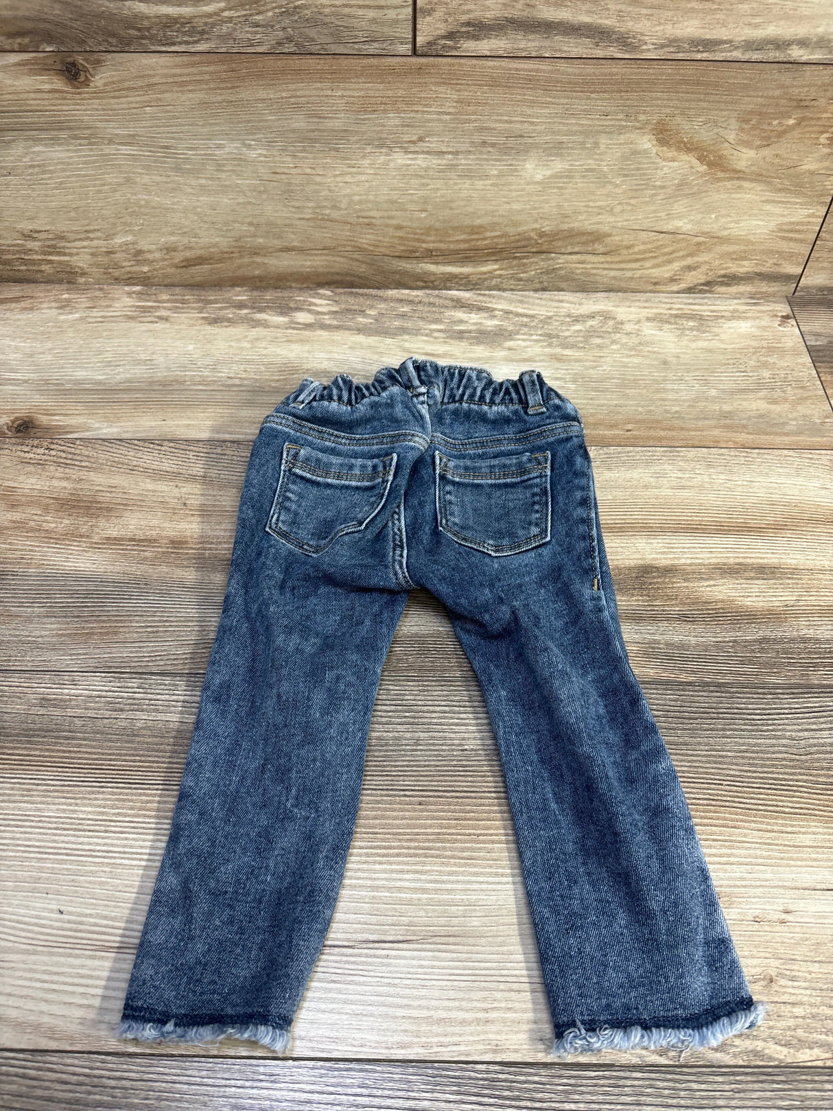 Back view of Old Navy jegging jeans blue size 18-24M with elastic ruffled waistband, back pockets and frayed hems on wood
