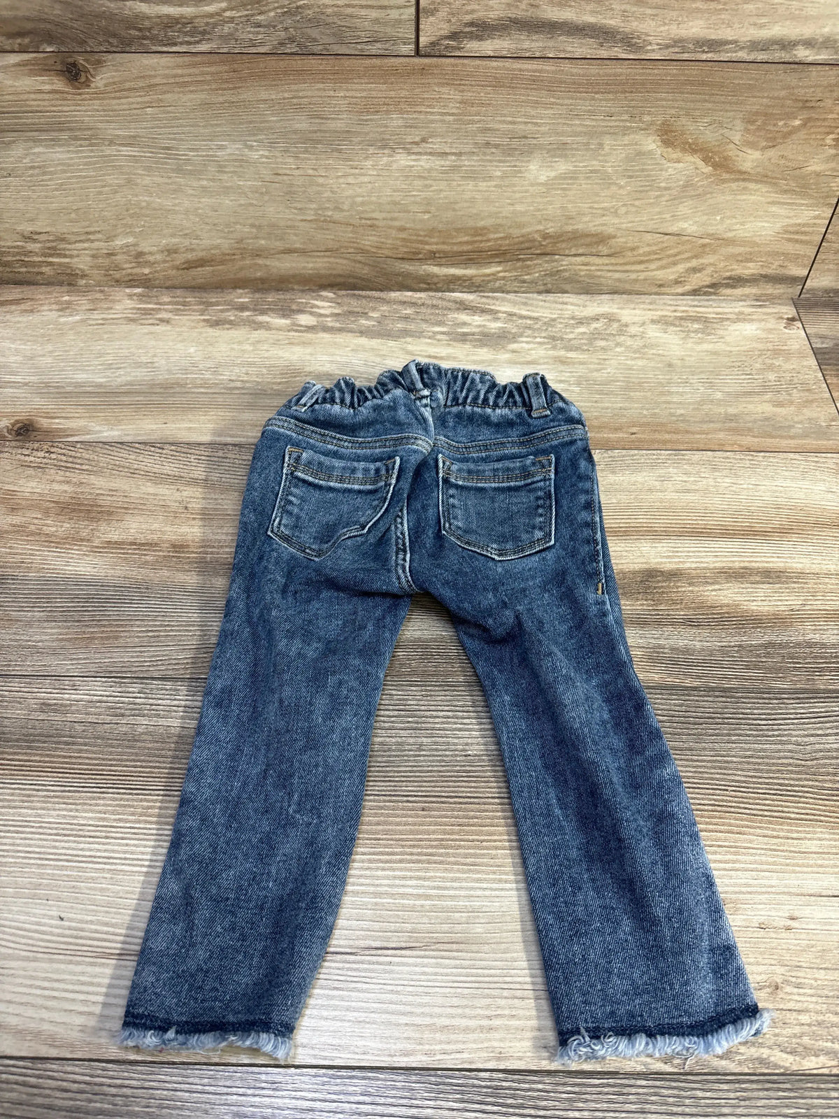 Back view of Old Navy jegging jeans blue size 18-24M with elastic ruffled waistband, back pockets and frayed hems on wood
