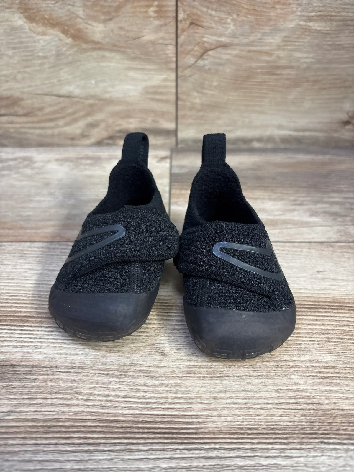 Front view of black Nike Swoosh 1 TD sneakers showcasing hook-and-loop straps and pull tabs on a wood-look floor