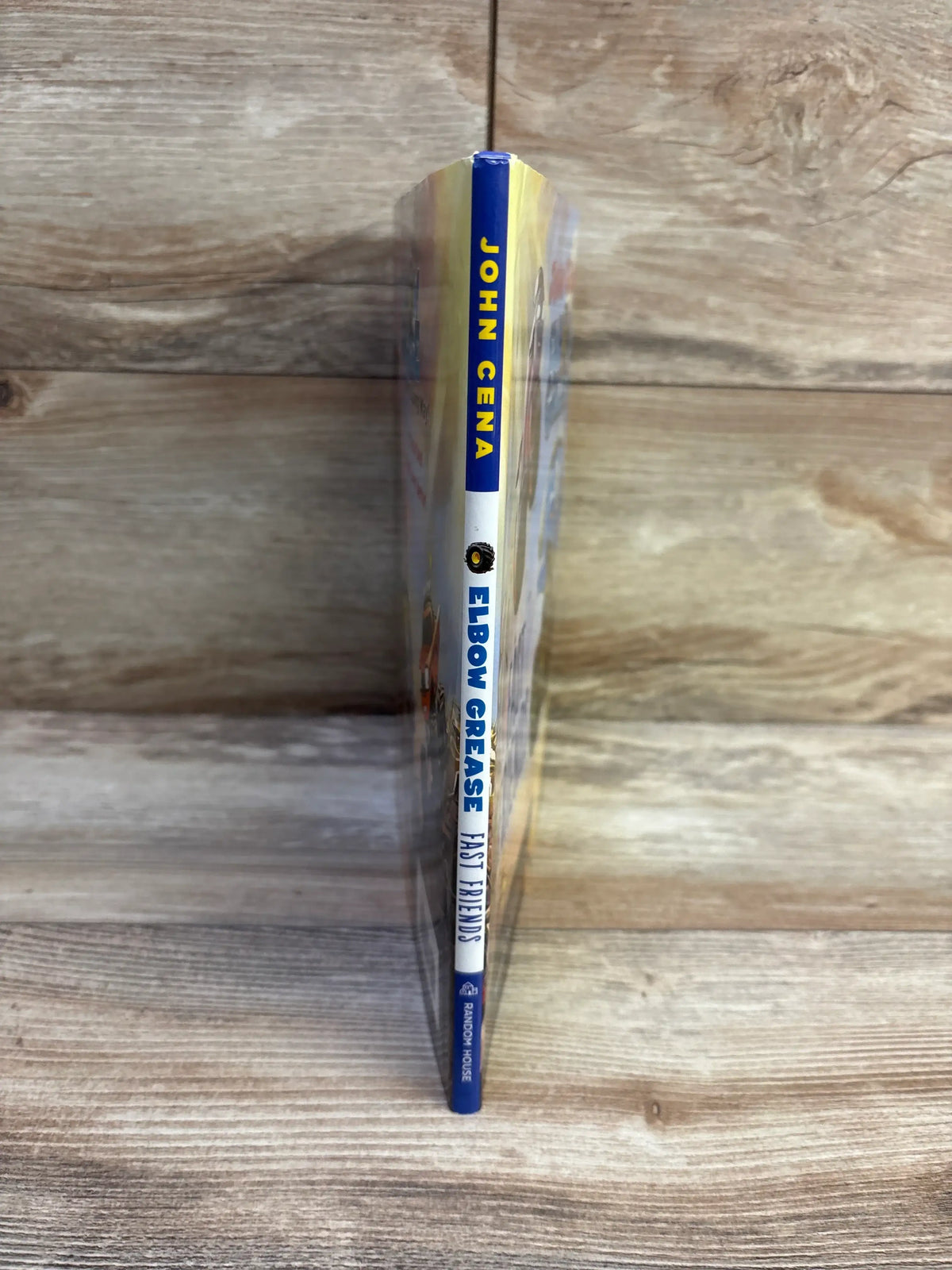 Spine view of John Cena Elbow Grease: Fast Friends hardcover showcasing spine title and publisher logo on wood background