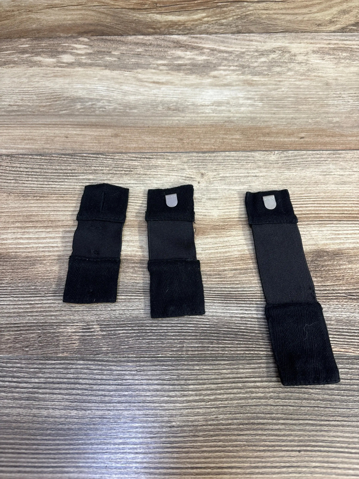 Top down view of three black belly belt strap extenders showing two short and one long strap with snap tabs on wood background