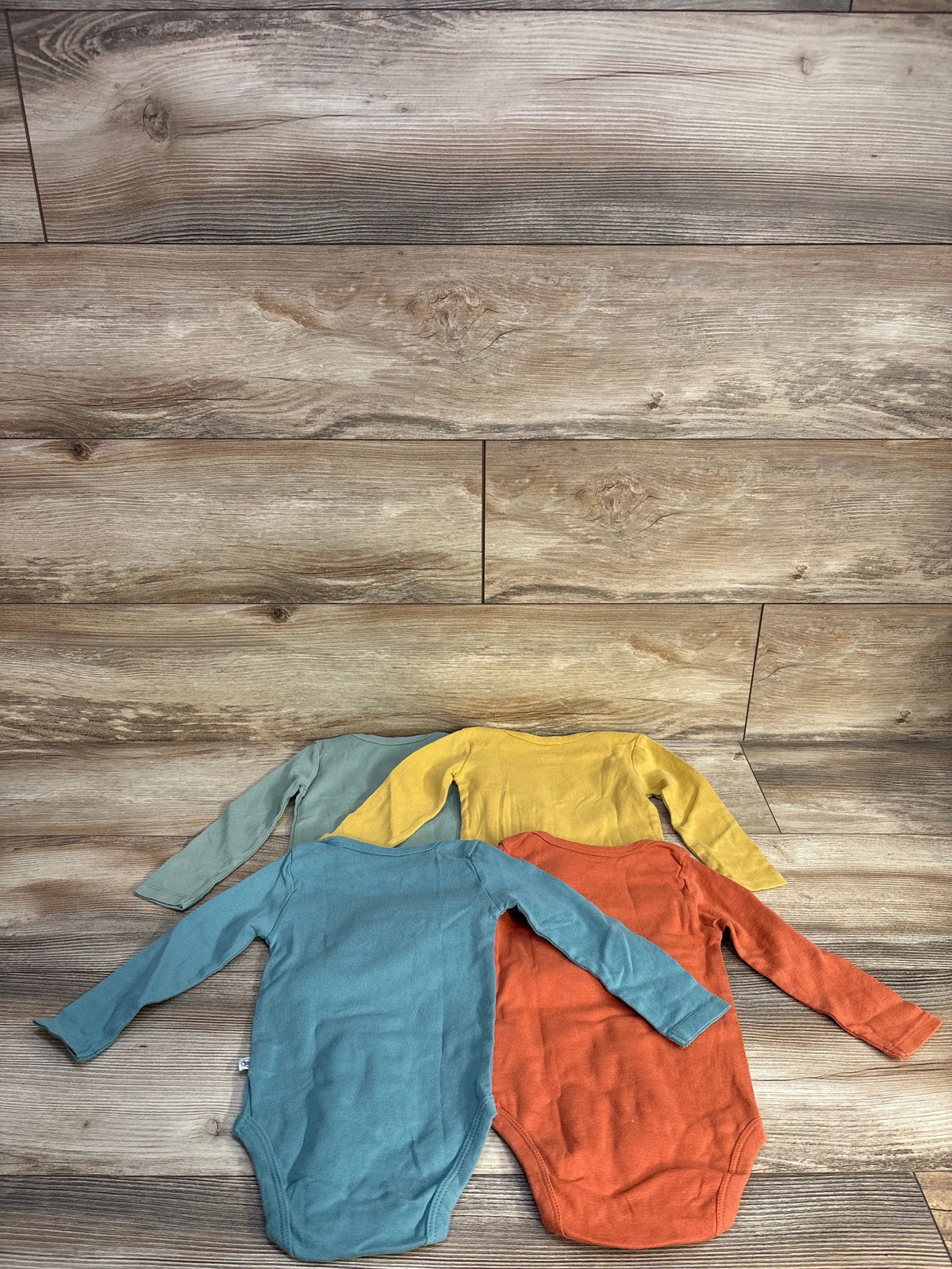 Top view of Honest Baby four-piece long sleeve bodysuit set in yellow, blue, orange, and blue-green on a wood-look background