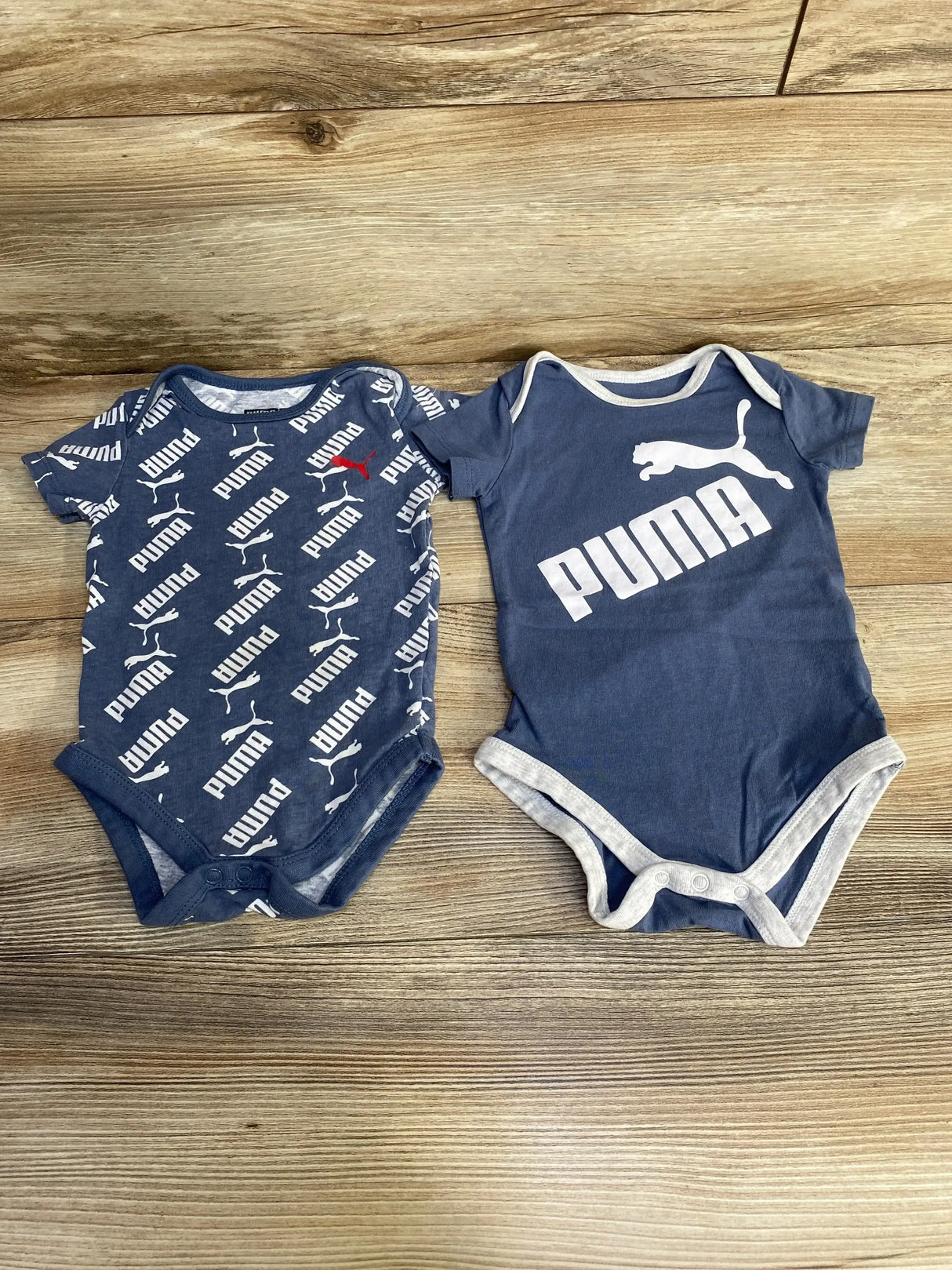 Top view of two blue Puma infant bodysuits: left all-over Puma logo print, right large Puma wordmark, on wood background