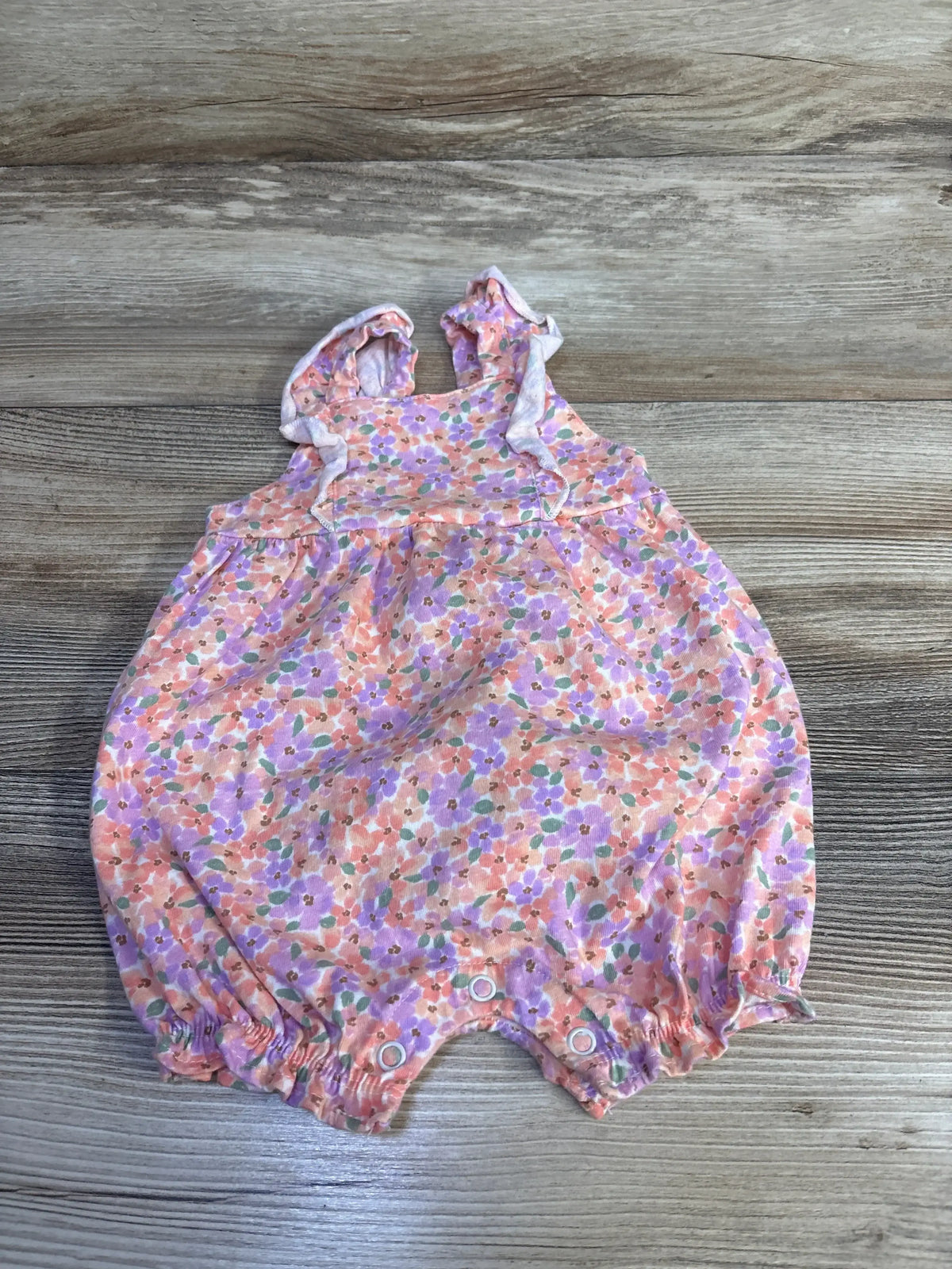 Flat lay of Just One You pink floral tank shortie romper size 3m showcasing ruffle straps and snap closure on wood plank floor