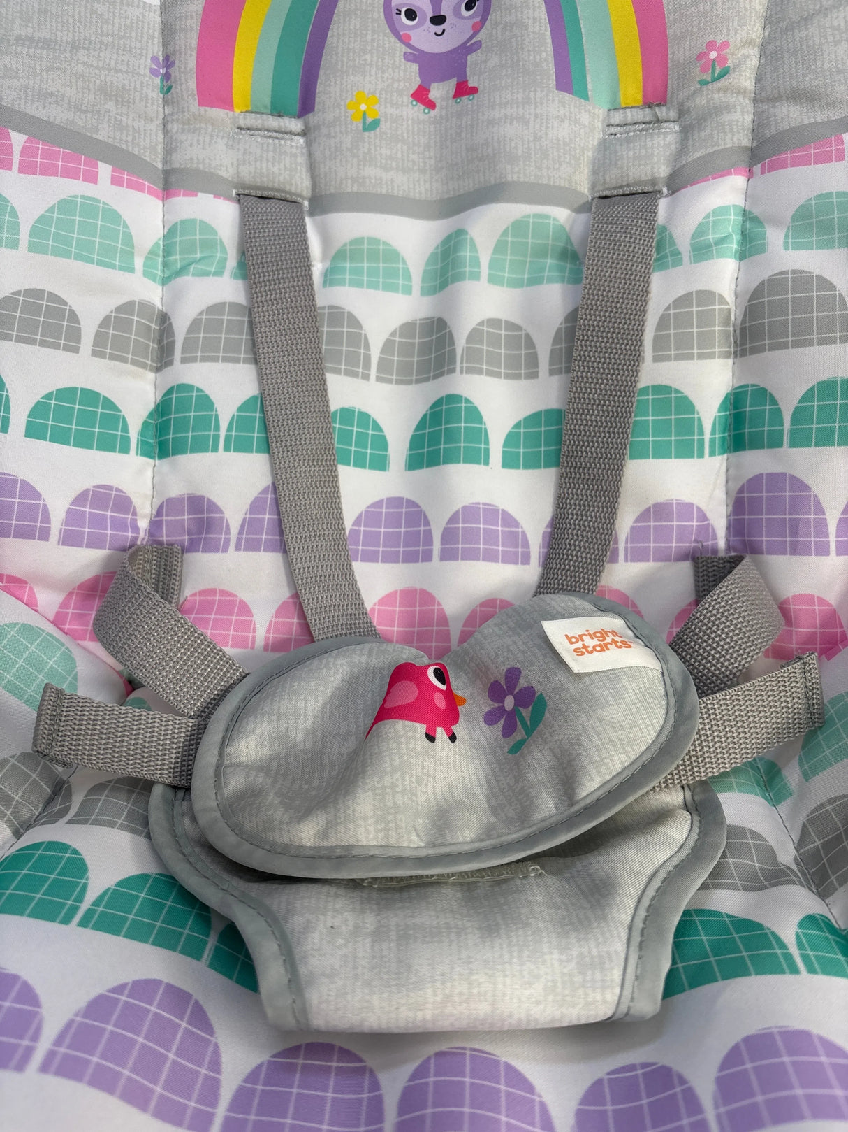 Close-up of Bright Starts Rosy Rainbow infant-to-toddler rocker harness showcasing padded crotch cover and gray safety straps with rainbow-arch patterned seat