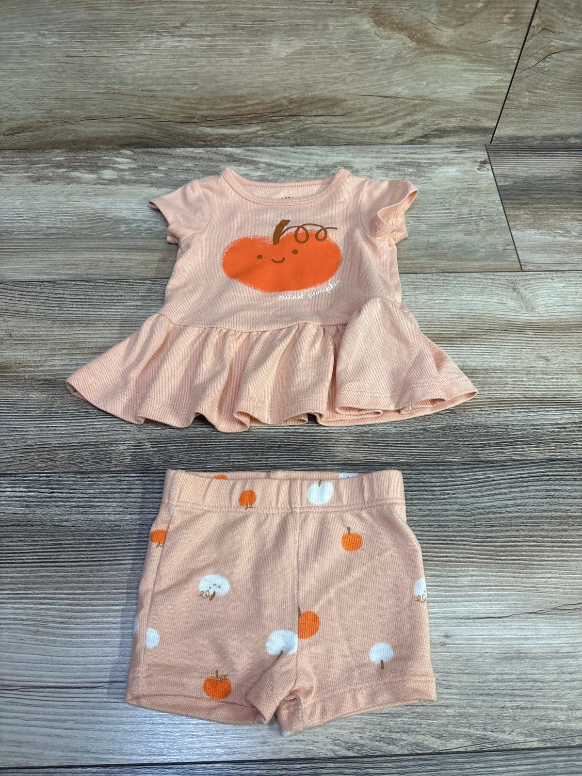 Top-down view of Cat and Jack peplum top and shorts, size 3 to 6 months, with pumpkin prints on wood-grain background