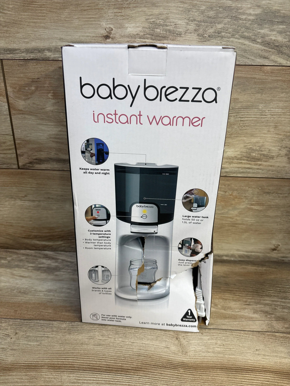 Front view of Baby Brezza Instant Warmer box showing warmer pouring water into bottle with torn lower-right corner on wood floor