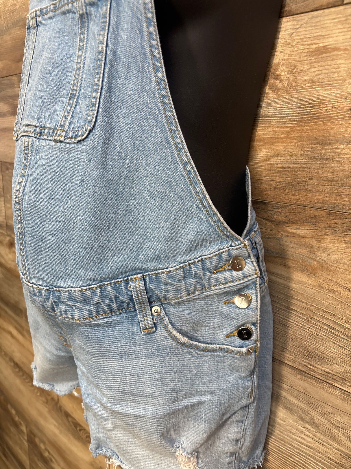 Side close-up of Isabel maternity denim shortalls showcasing side button closures, front bib pocket, and frayed hem on wood background
