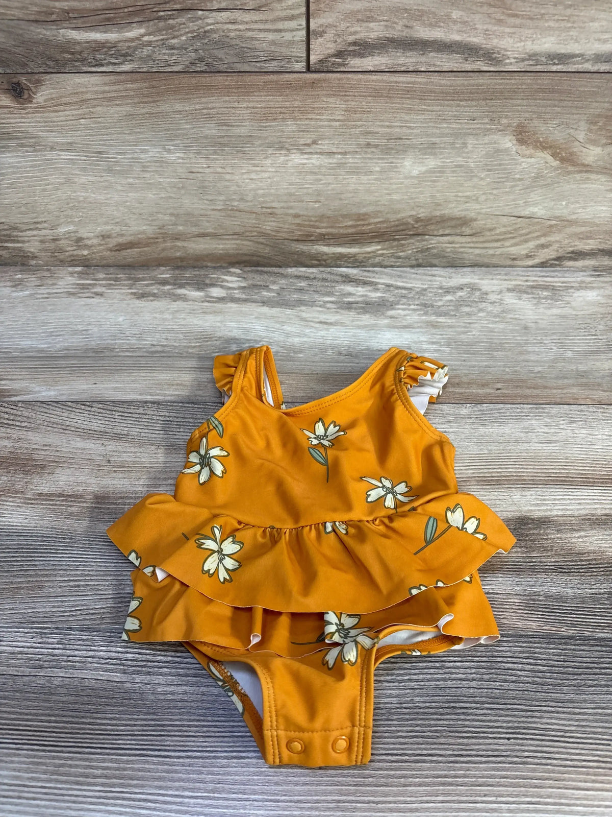 Top-down view of orange one-piece floral ruffle swimsuit with layered ruffles and snap crotch on wood background, size 6M