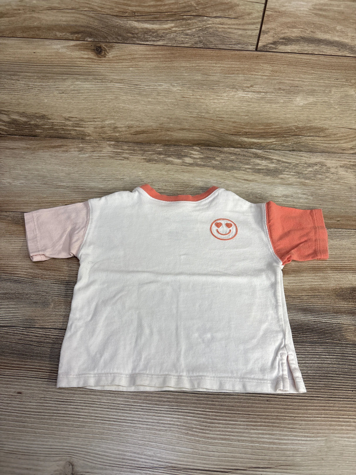 Back view of white kids t-shirt with pink and orange sleeves and smiley face embroidery on wooden floor