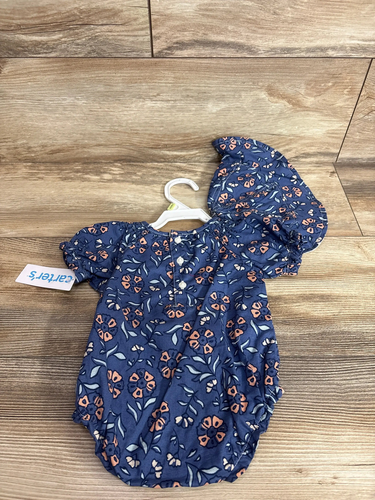 Top-down view of Carter's navy floral bubble romper with bucket hat showing puff sleeves and front buttons on wood floor
