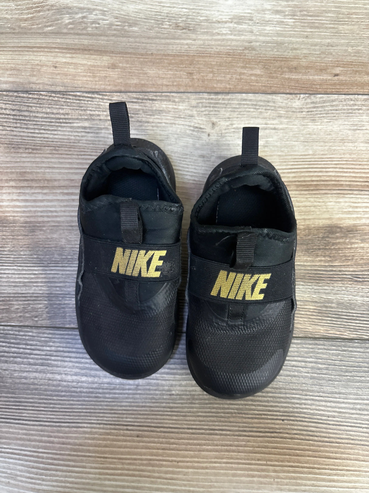 Overhead view of Nike Flex Runner toddler sneakers size 8C showing black slip on design with gold NIKE strap on wood floor