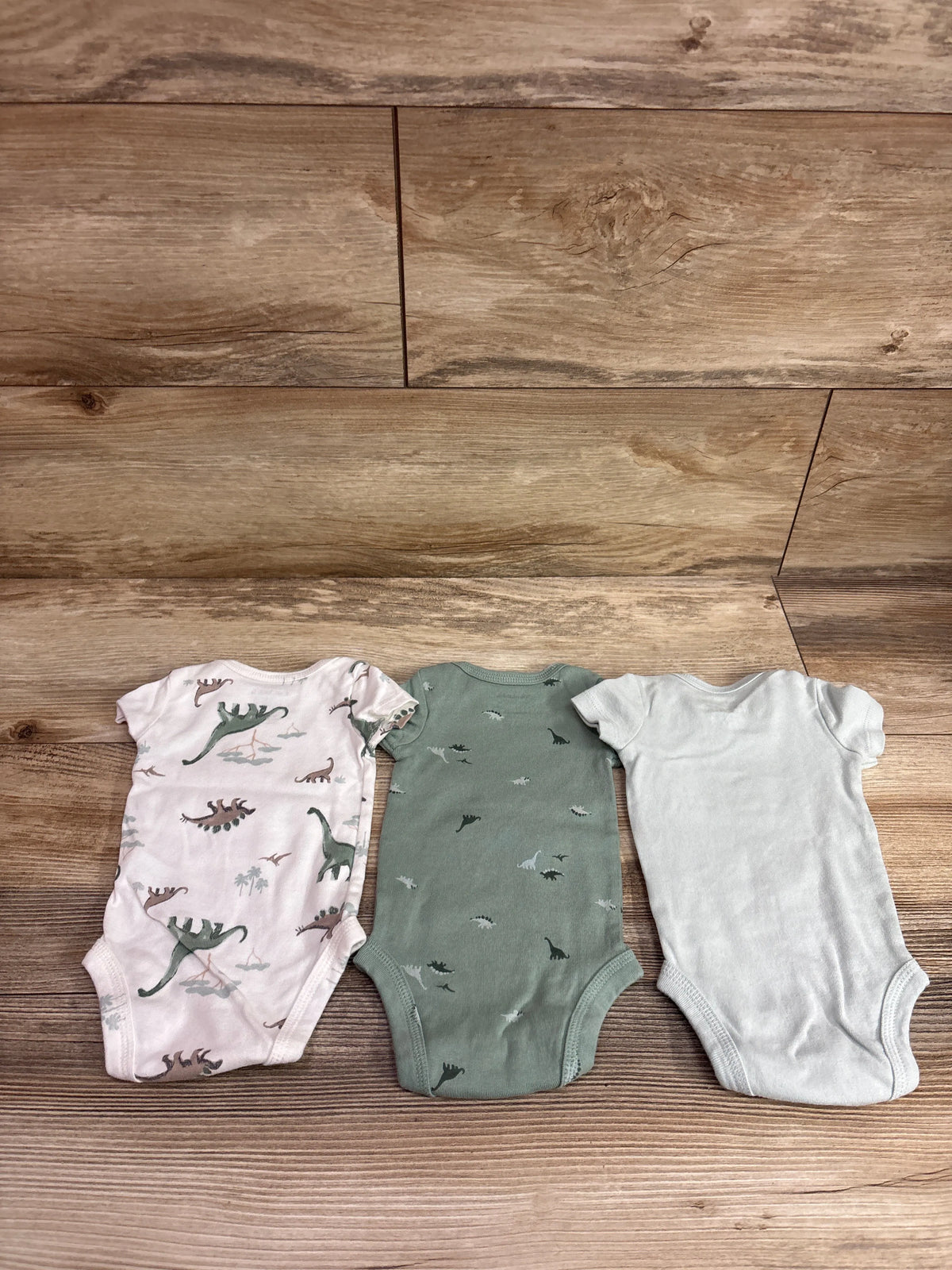 Top view of three Just One You newborn bodysuits with dinosaur prints and solid white design on a wood background
