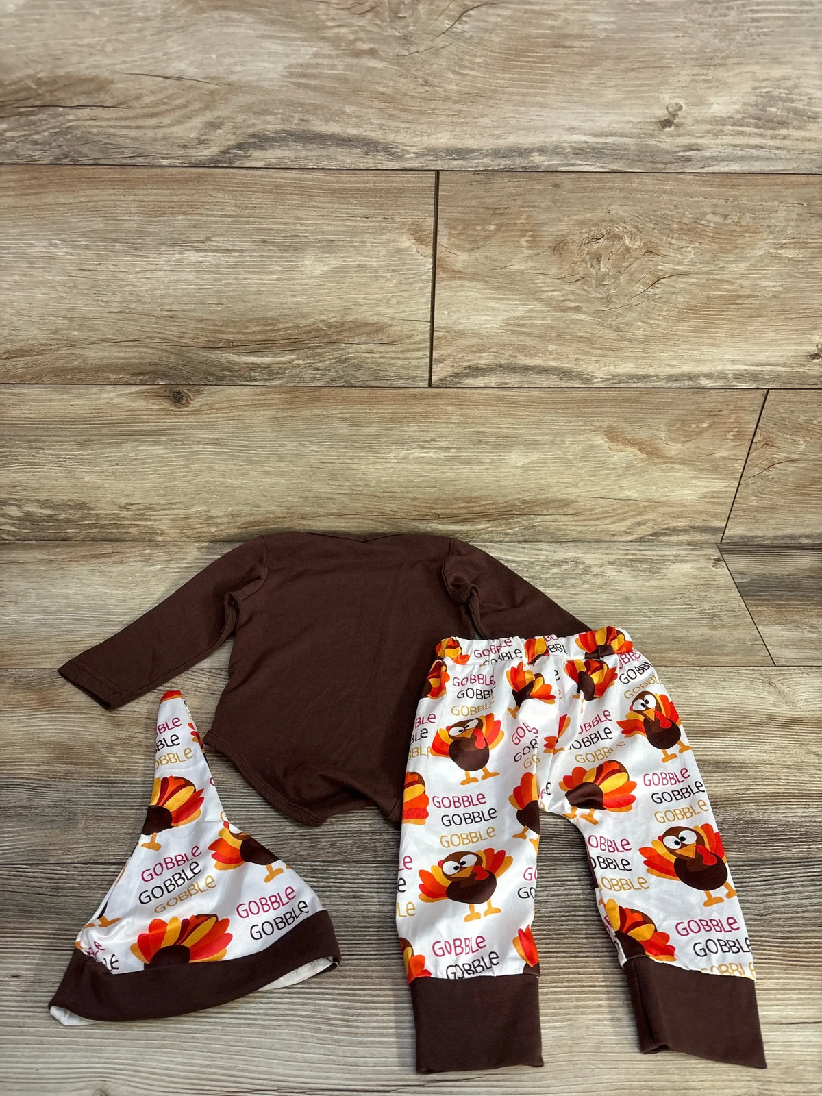 Baby fall outfit with brown long sleeve onesie, Thanksgiving turkey print pants and matching hat on wood floor