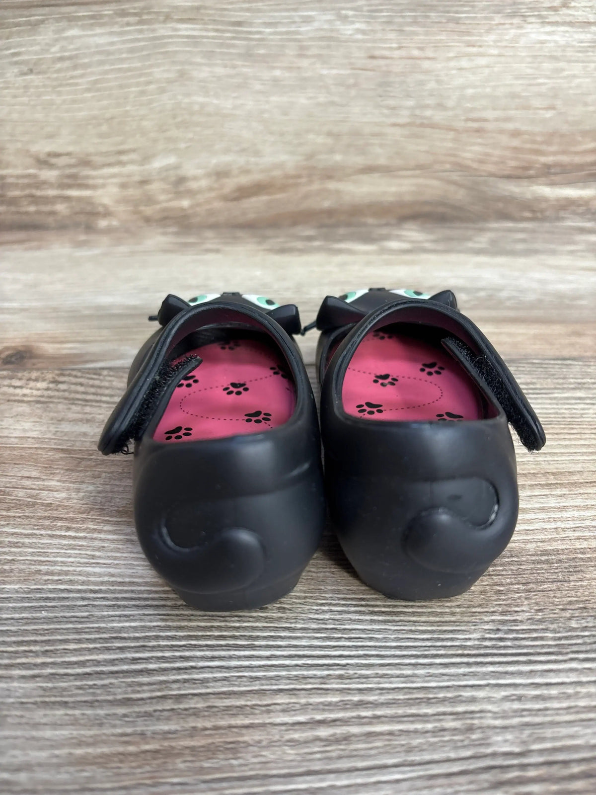 Back view of children's black shoes with cat face design and pink insoles with paw prints on wooden floor