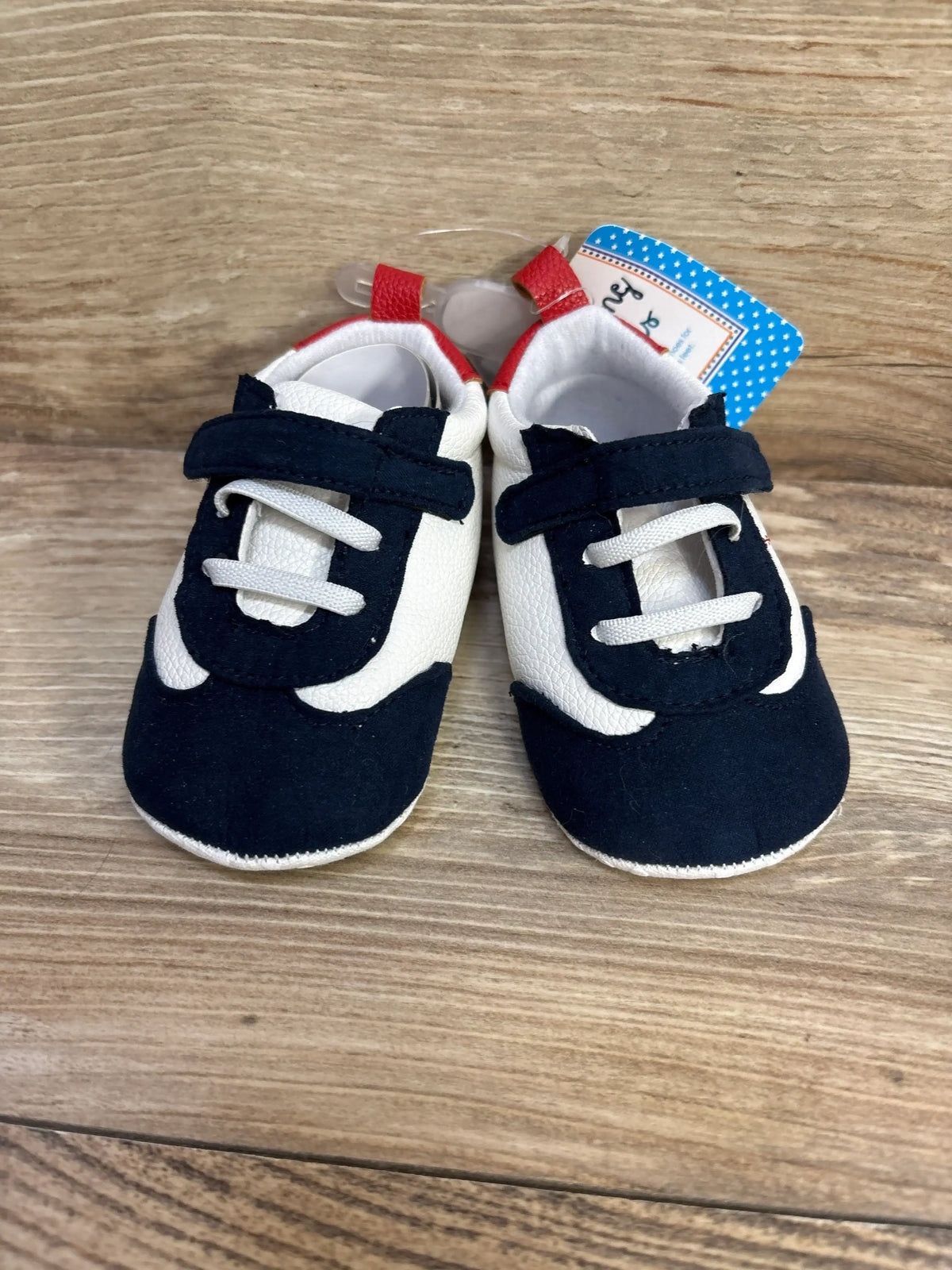 Front view of baby baseball-style shoes in navy and white with Velcro strap, elastic laces, red heel tabs on wood background