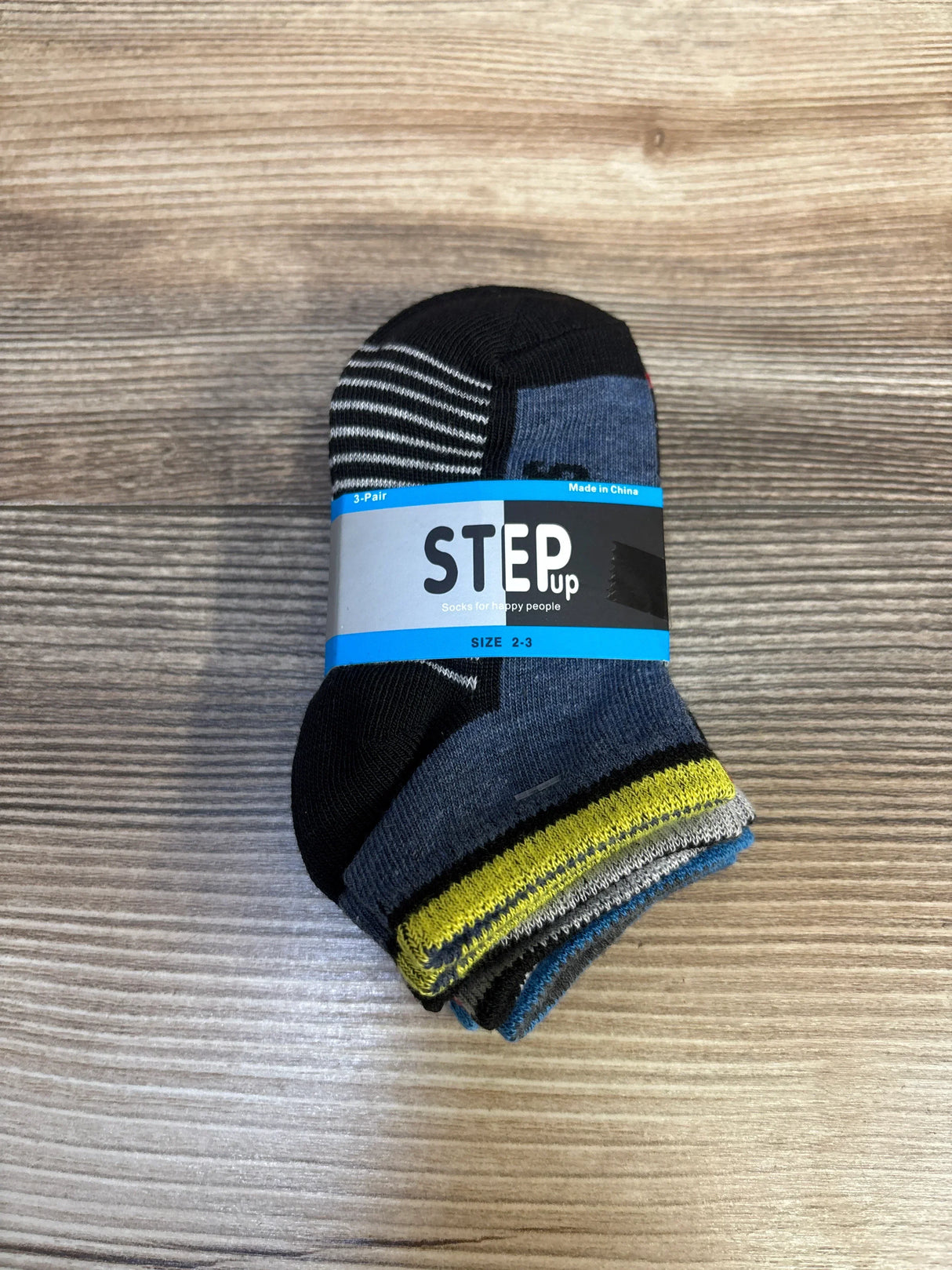 Top-down view of new STEP up 3-pack pattern socks size 2-3 with black grey yellow and blue striped designs in label band on wood plank floor