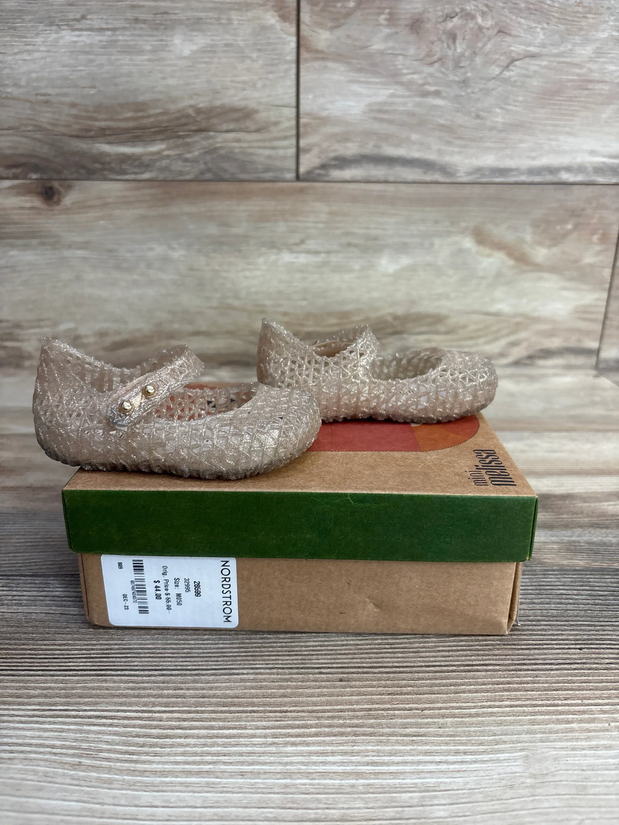 Side view of Mini Melissa Campana jelly flats in gold showing glitter finish and Mary Jane strap on cardboard box