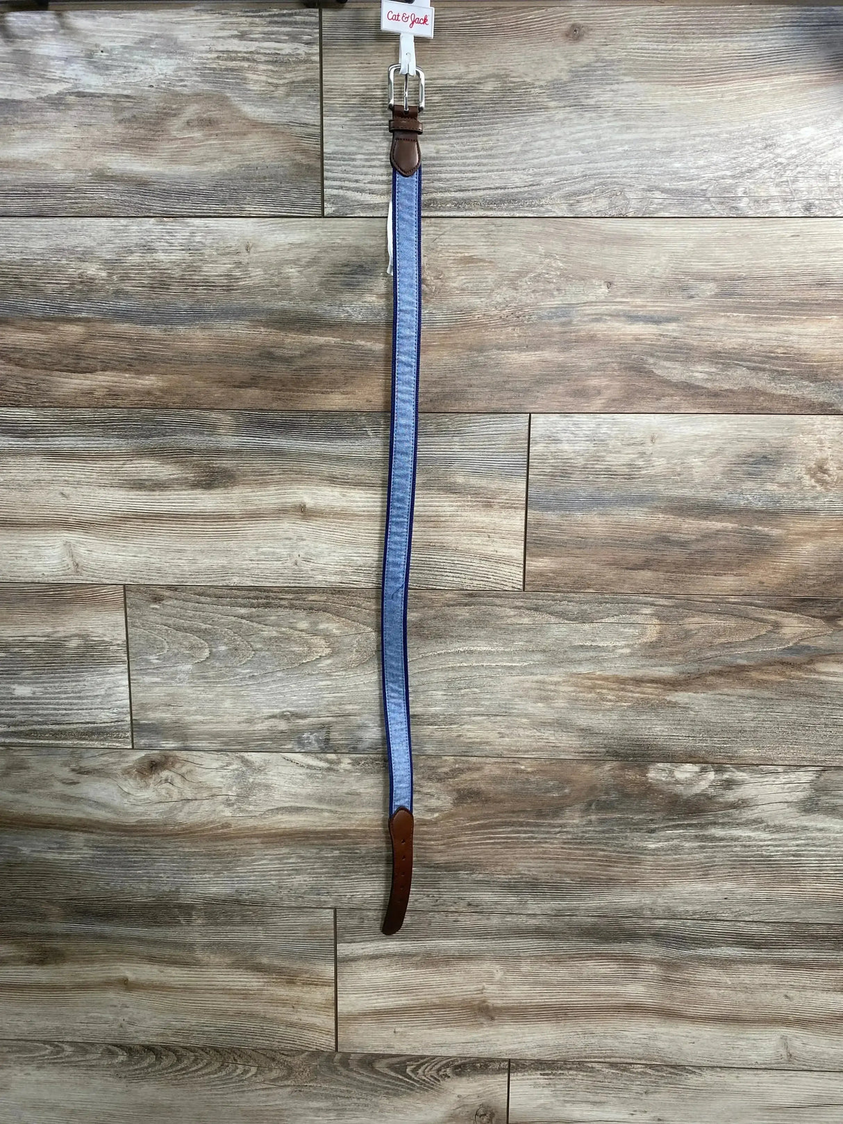 Front view of Cat and Jack chambray belt in blue with brown leather tipping and metal buckle with hangtag on wood plank floor