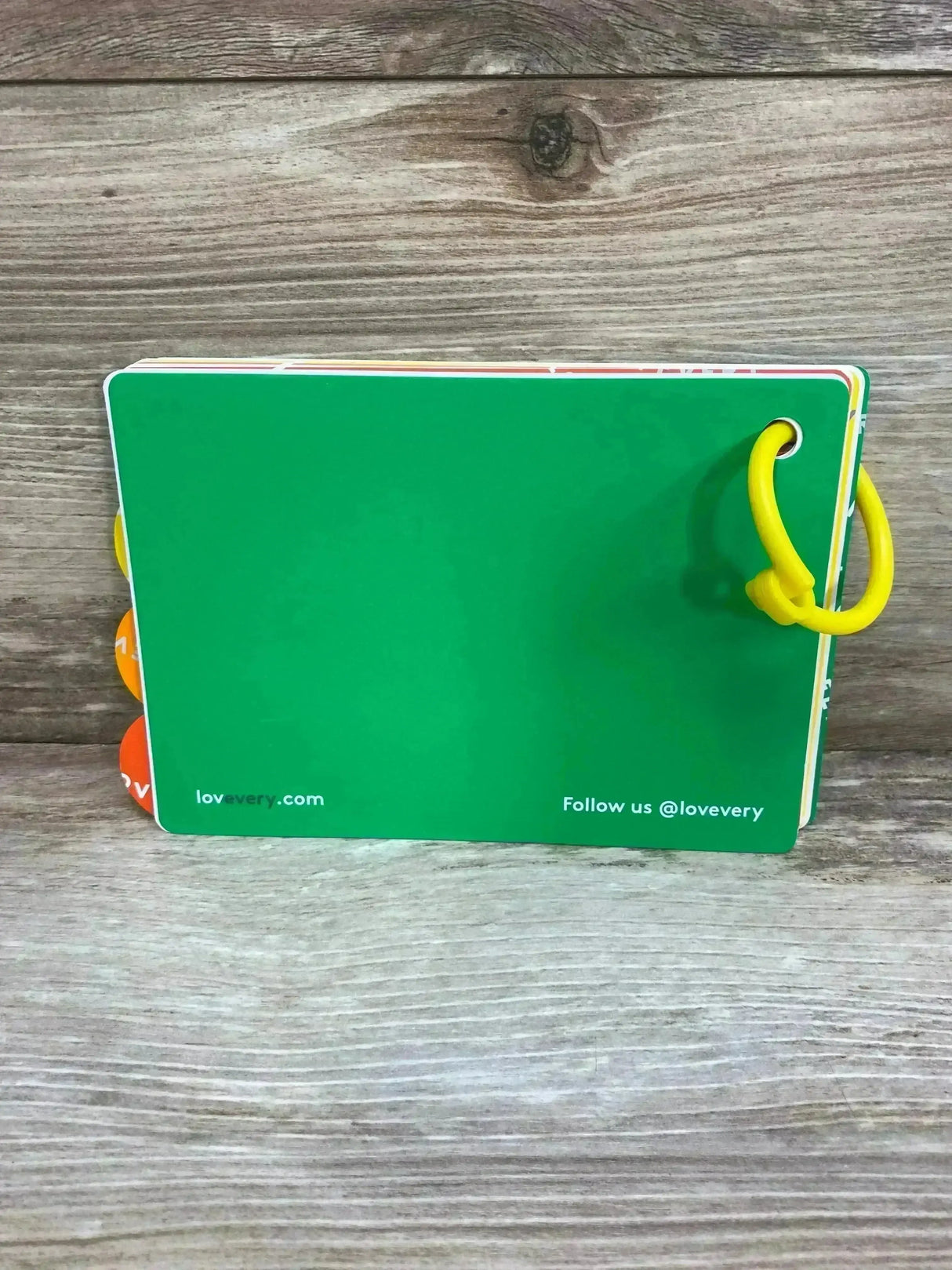 Back view of Lovevery The Thinkers Play Guide 11-12m showcasing green back cover and yellow plastic ring with wood plank background
