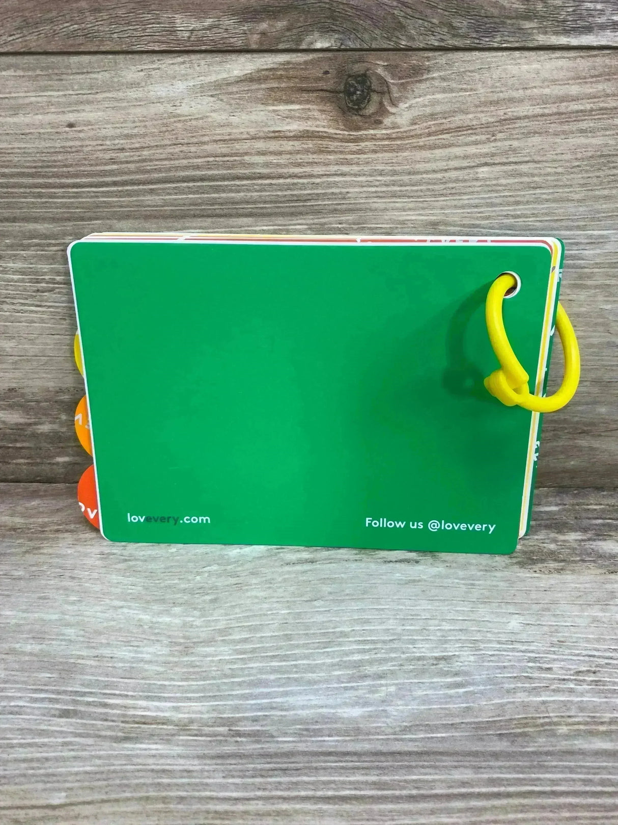 Back view of Lovevery The Thinkers Play Guide 11-12m showcasing green back cover and yellow plastic ring with wood plank background