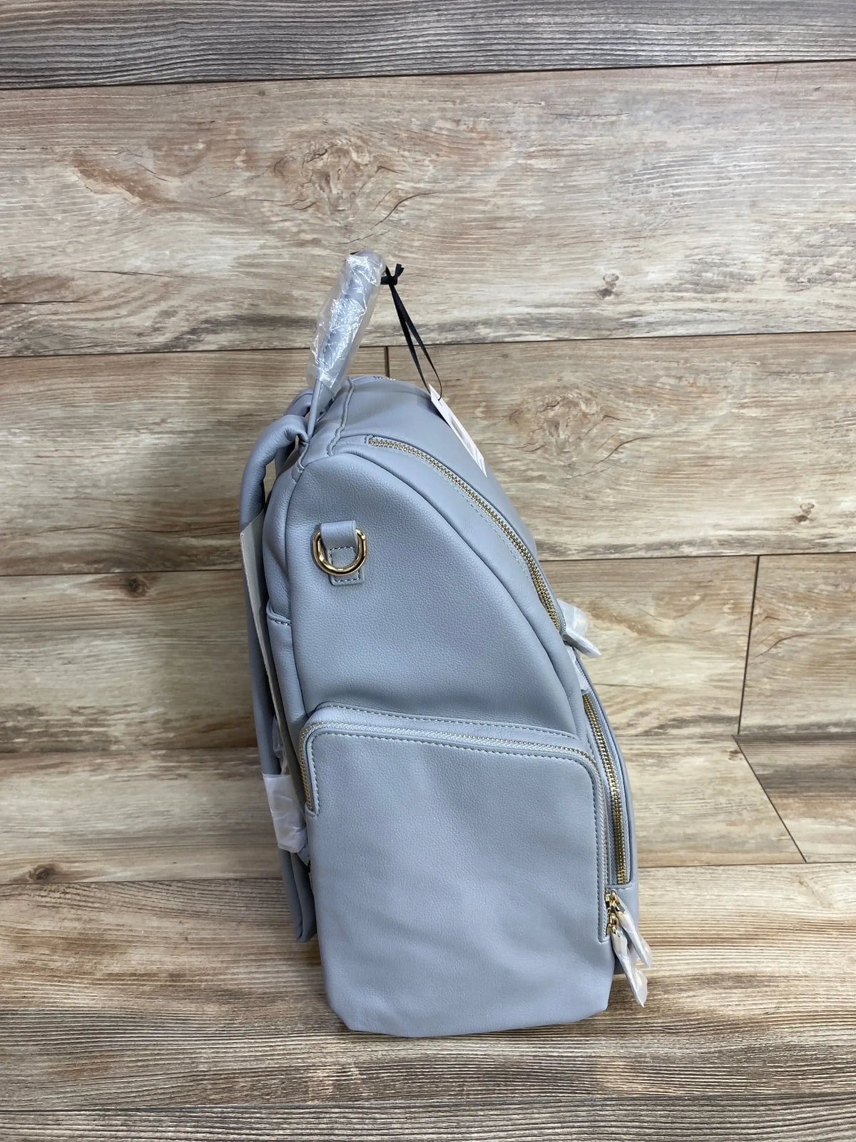 Side view of KeriKit Amber Apple diaper backpack in grey faux leather showing gold zippers, front pocket, D-ring and tags on wood background