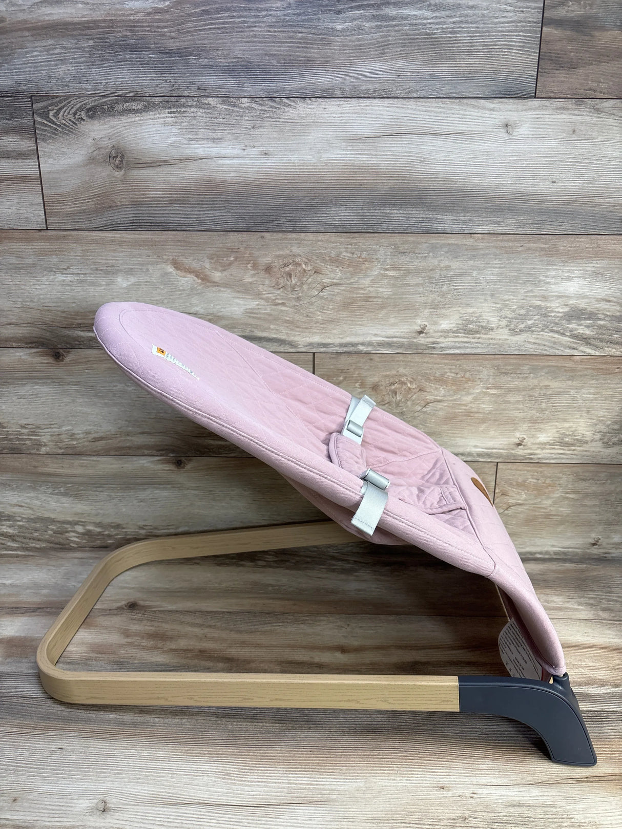 Side view of ANGELBLISS baby bouncer in pink showing quilted seat, harness, and light wood base on a wood plank background