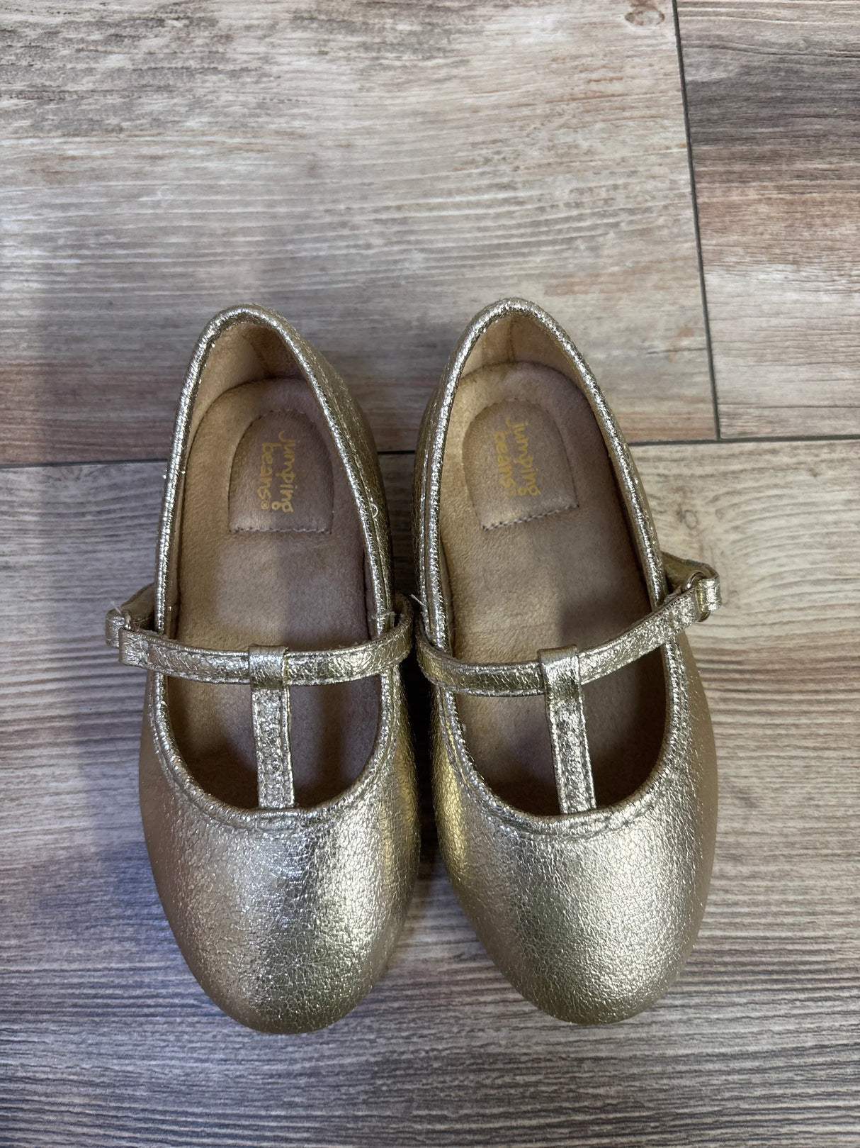 Top-down view of Jumping Beans Susie T-strap ballet flats showcasing metallic gold finish and buckle on wood floor