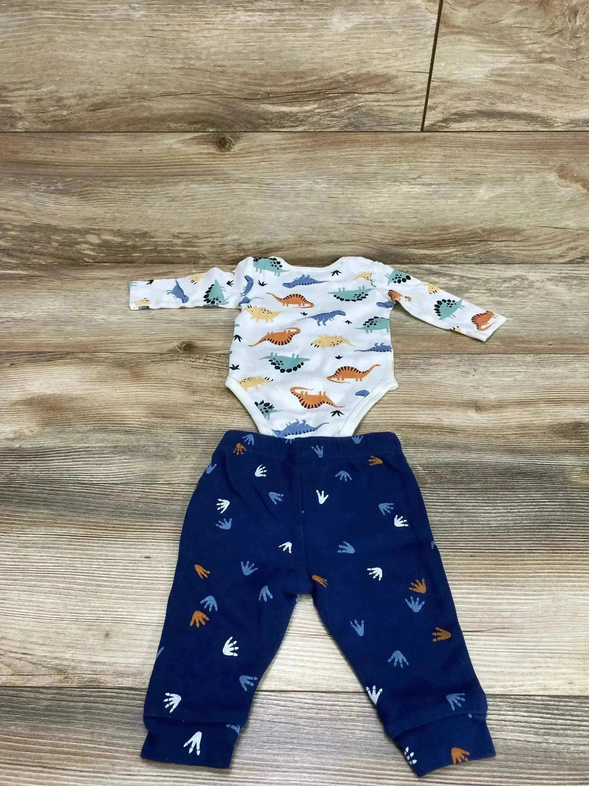 Flat lay of Pekkle 2-piece set: white dinosaur-print bodysuit and navy footprint-print pants on wood background