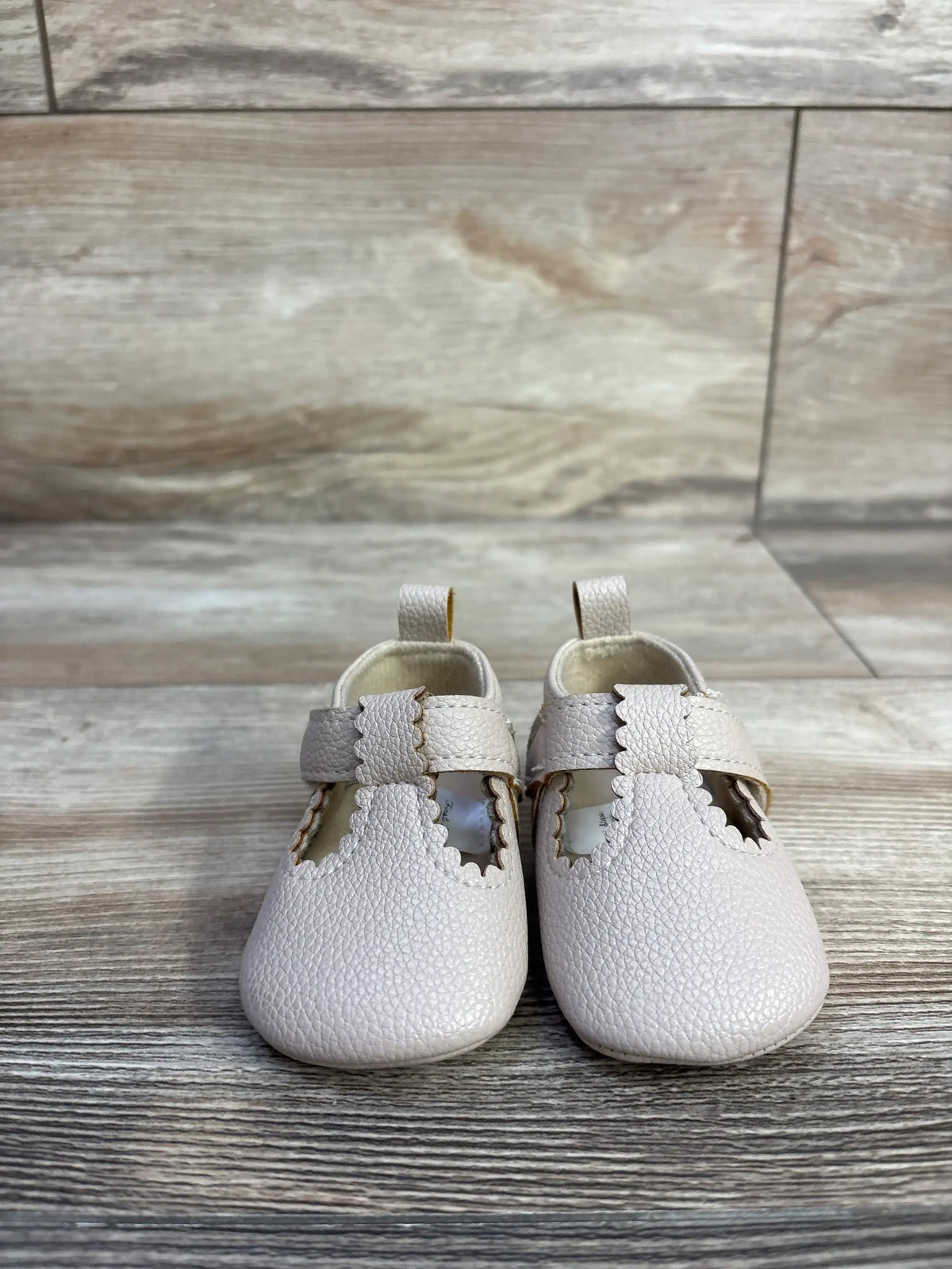 Front view of First Steps soft-sole baby shoes showcasing scalloped T-straps and pebbled beige leather on a wood-look floor