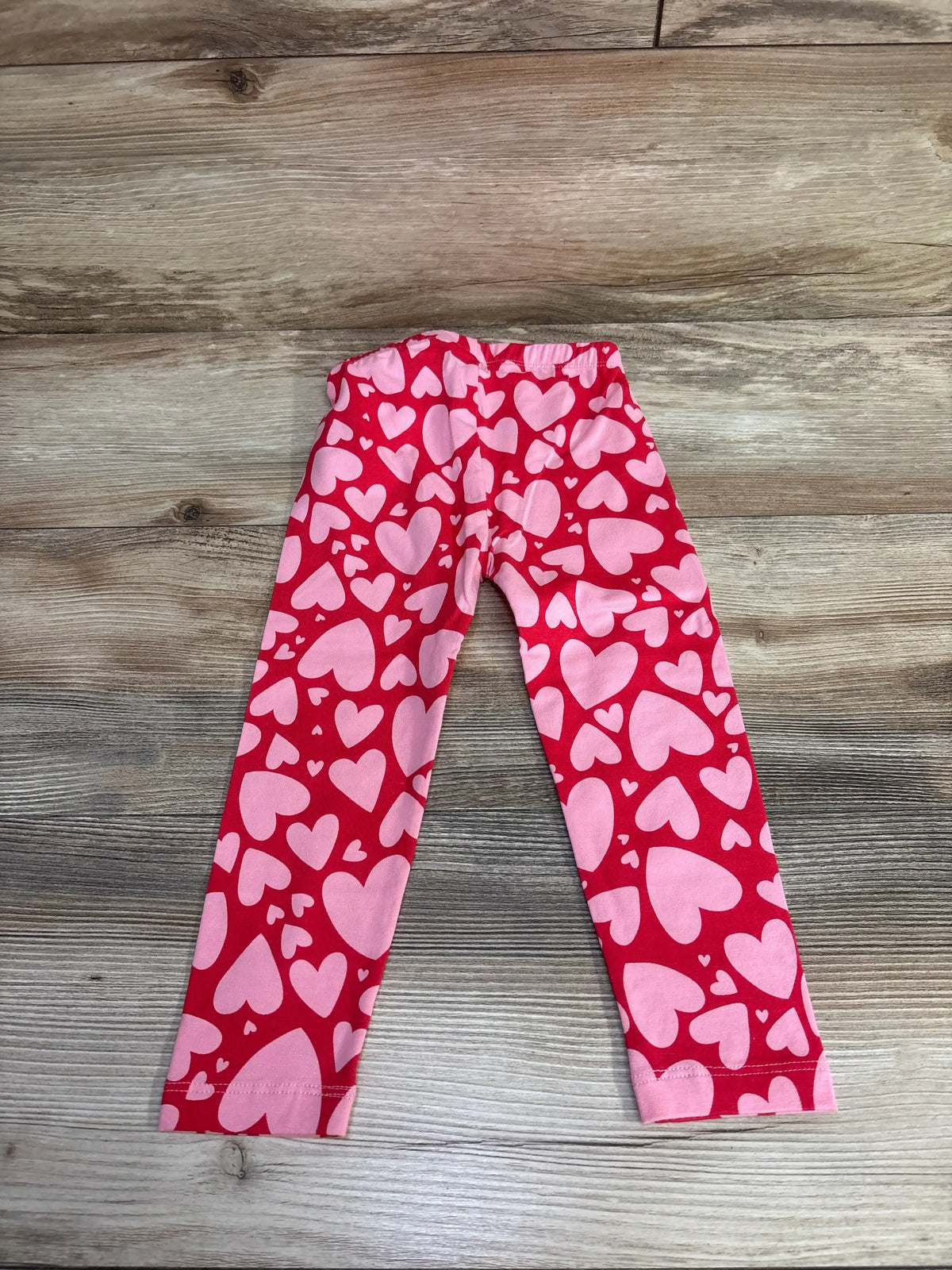Top-down view of Old Navy heart leggings in red, size 2T, showcasing pink hearts on a wood-grain background