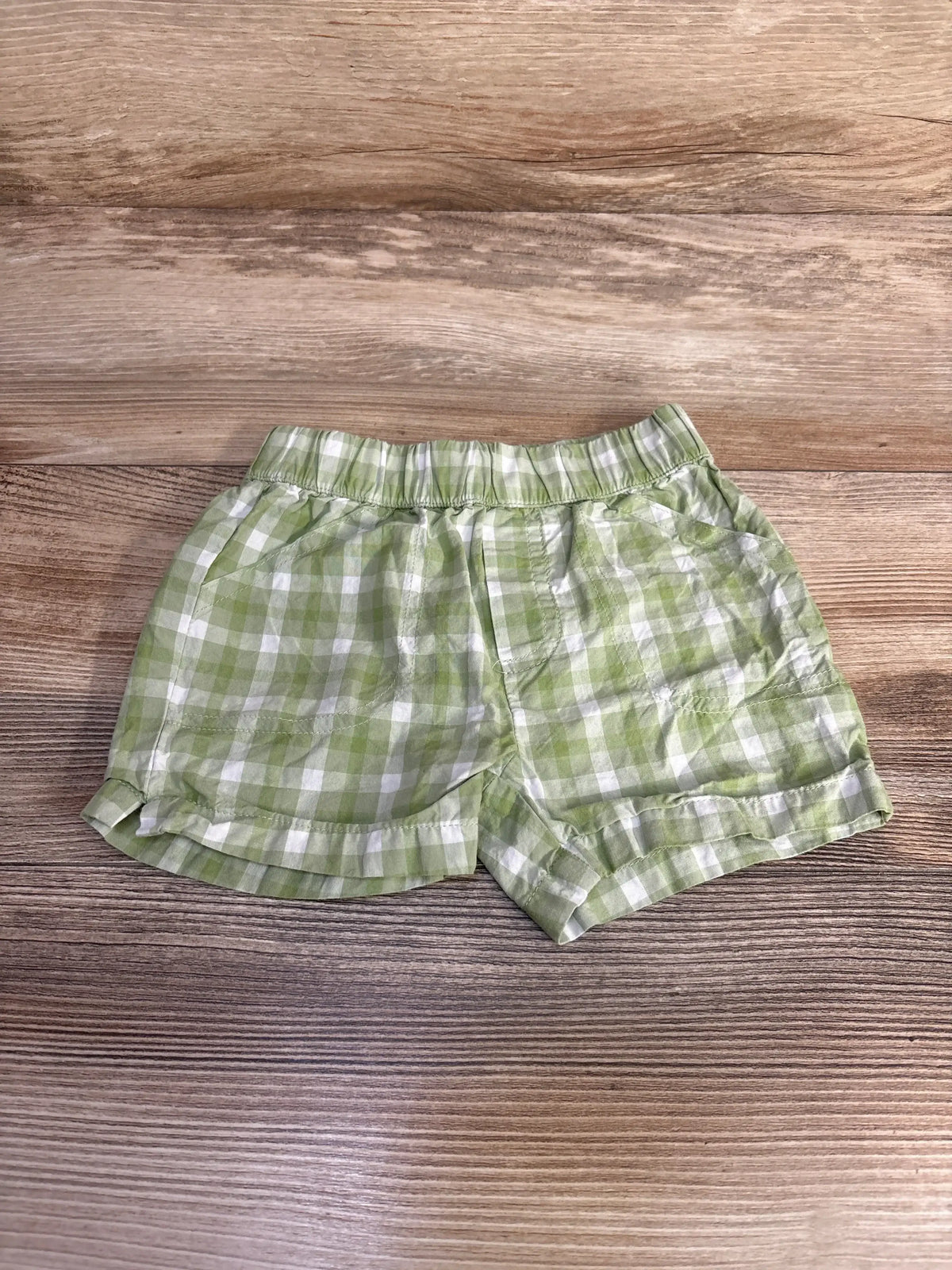 Top-down view of green and white gingham plaid toddler shorts showing elastic waistband and ruffle hem on wood background