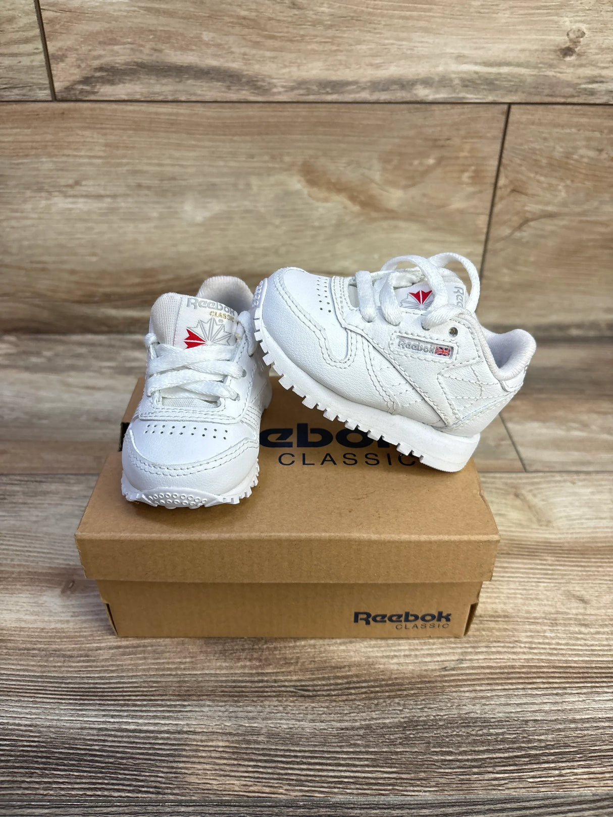 Angled front view of white Reebok Classic Leather infant sneakers showing leather upper, laces, logo on Reebok shoebox