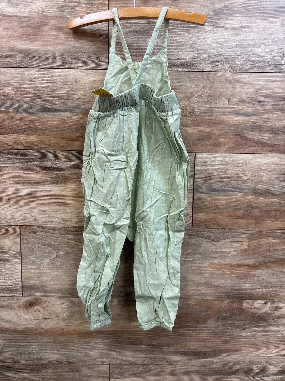 Front view of green toddler jumpsuit with crossed back straps, elastic gathered waist and relaxed pant legs on wood background, size 18 to 24 months