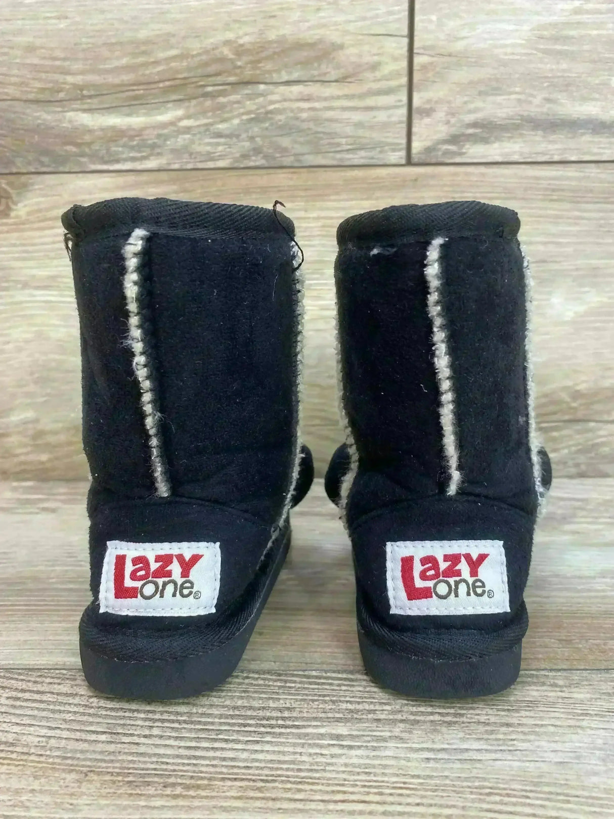 Rear view of black Lazy One kids moose slipper boots showing heel logo Lazy One and white faux fur seams on wood floor