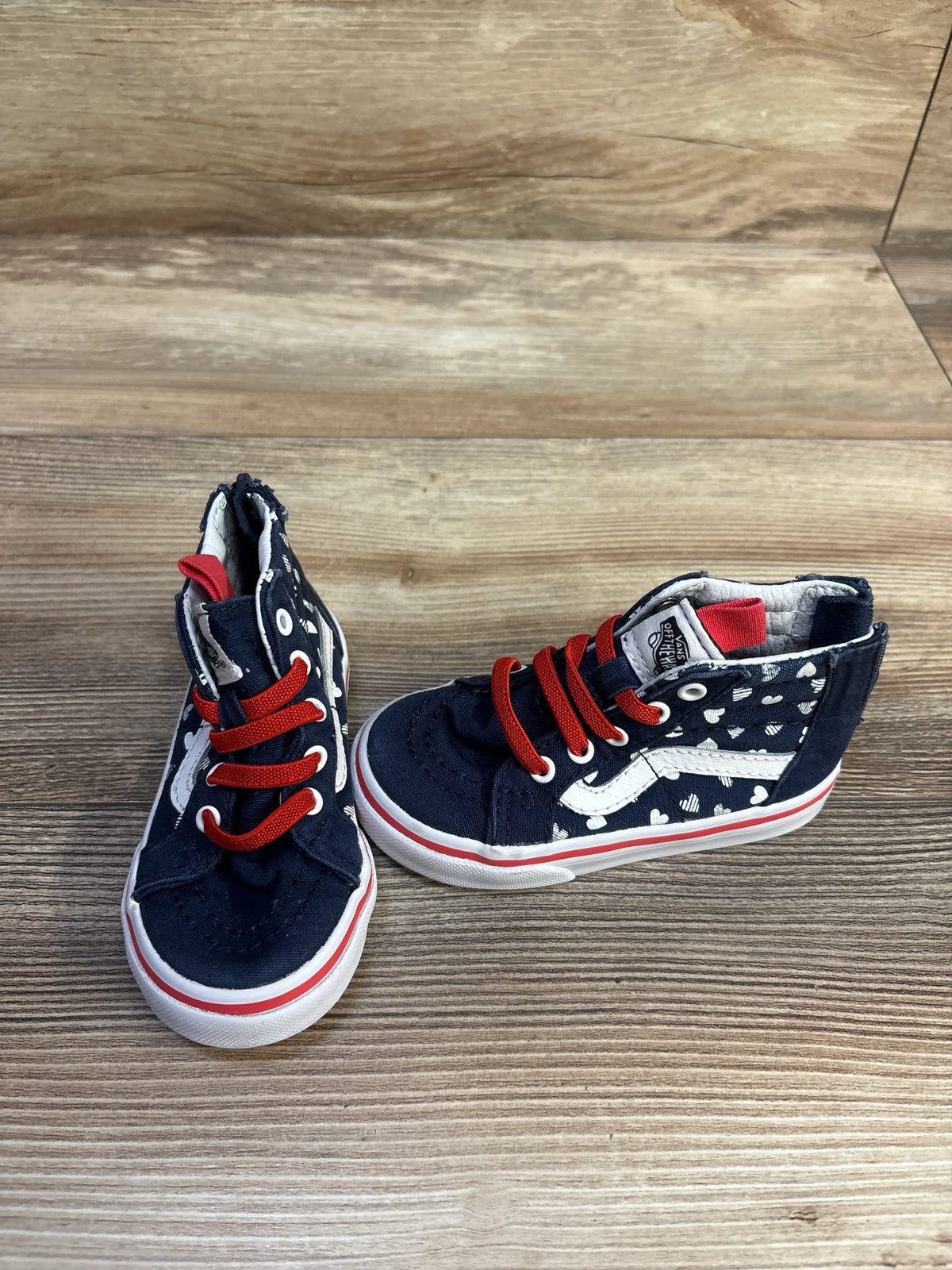 Three-quarter view of toddler Vans Sk8-Hi zip sneakers, navy with white hearts, red laces and side stripe on wood surface