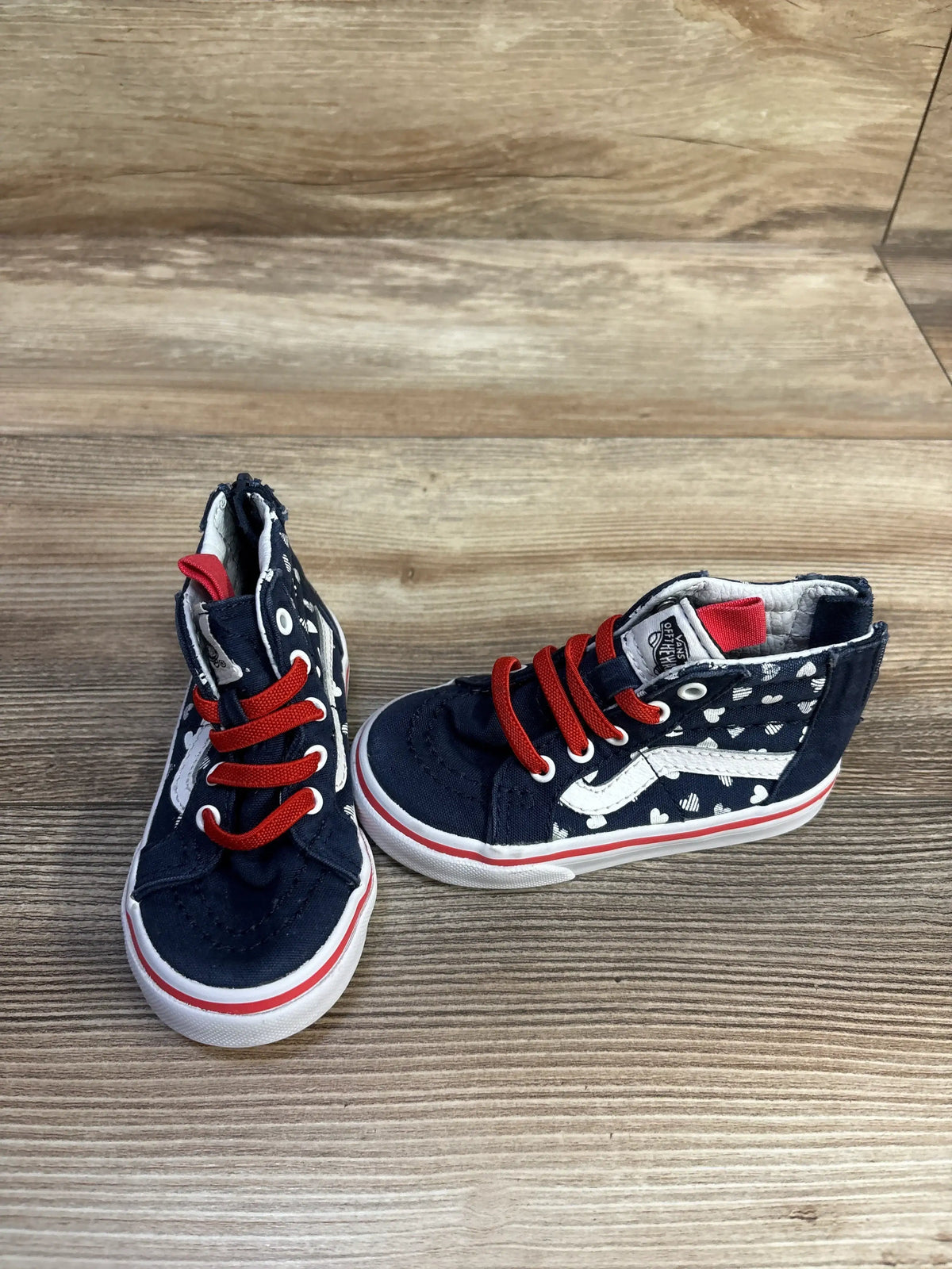 Three-quarter view of toddler Vans Sk8-Hi zip sneakers, navy with white hearts, red laces and side stripe on wood surface