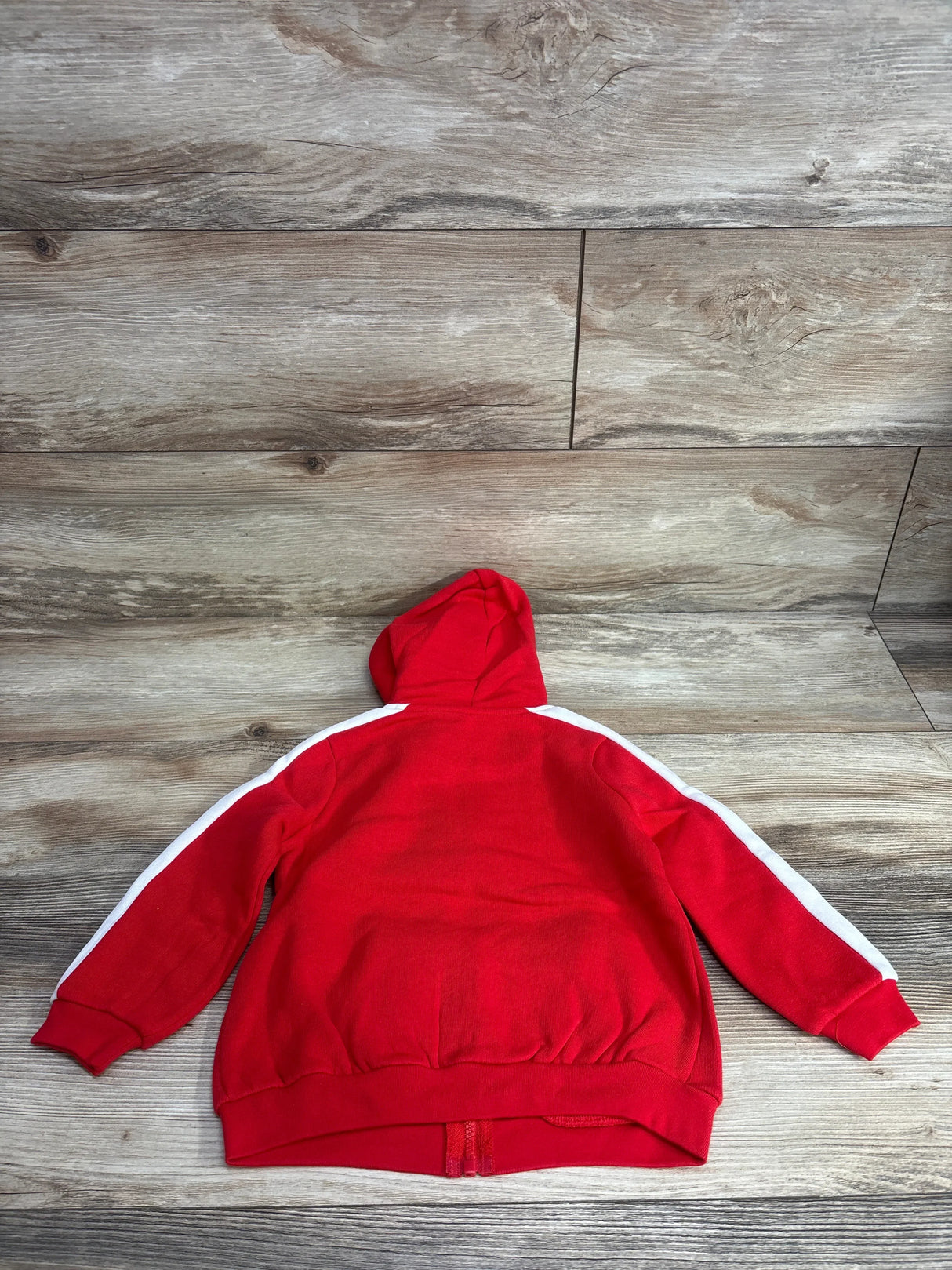 Red kids hoodie with white stripes on sleeves, laid flat on wooden surface