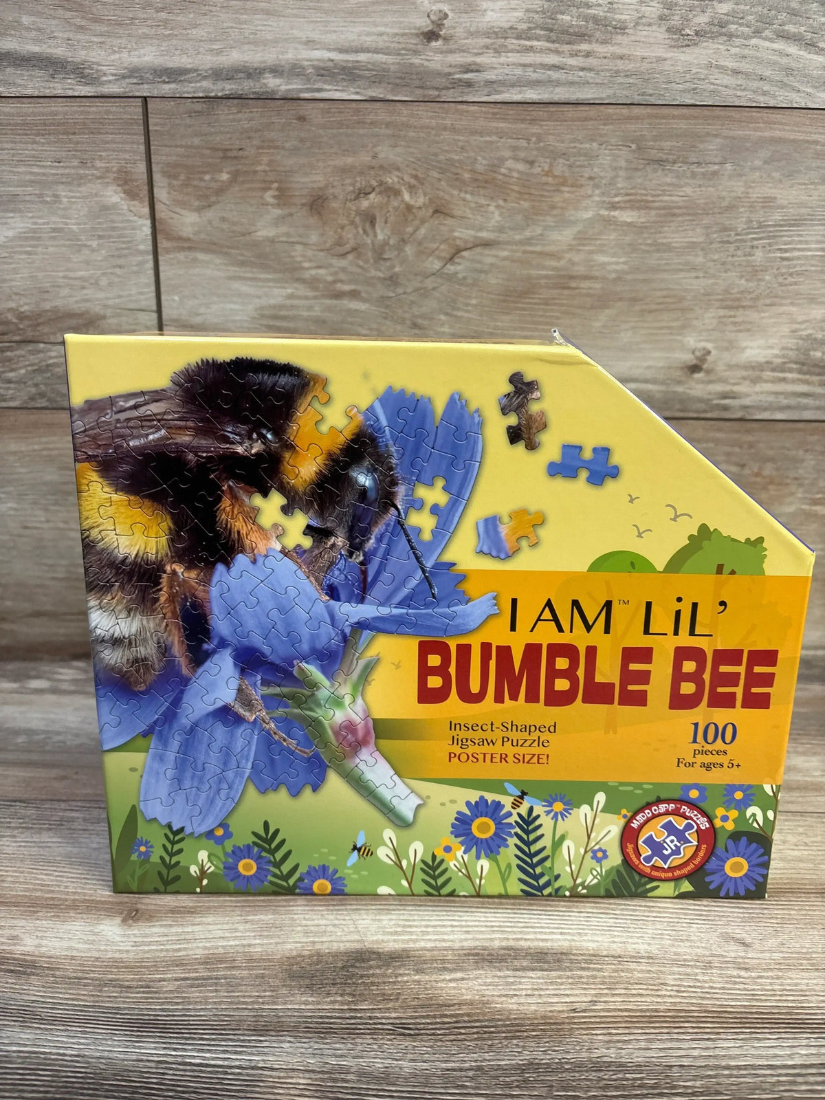 Front-angled view of Madd CAPP Lil' Bumble Bee 100-piece puzzle box with bumblebee artwork and insect shape on wooden surface