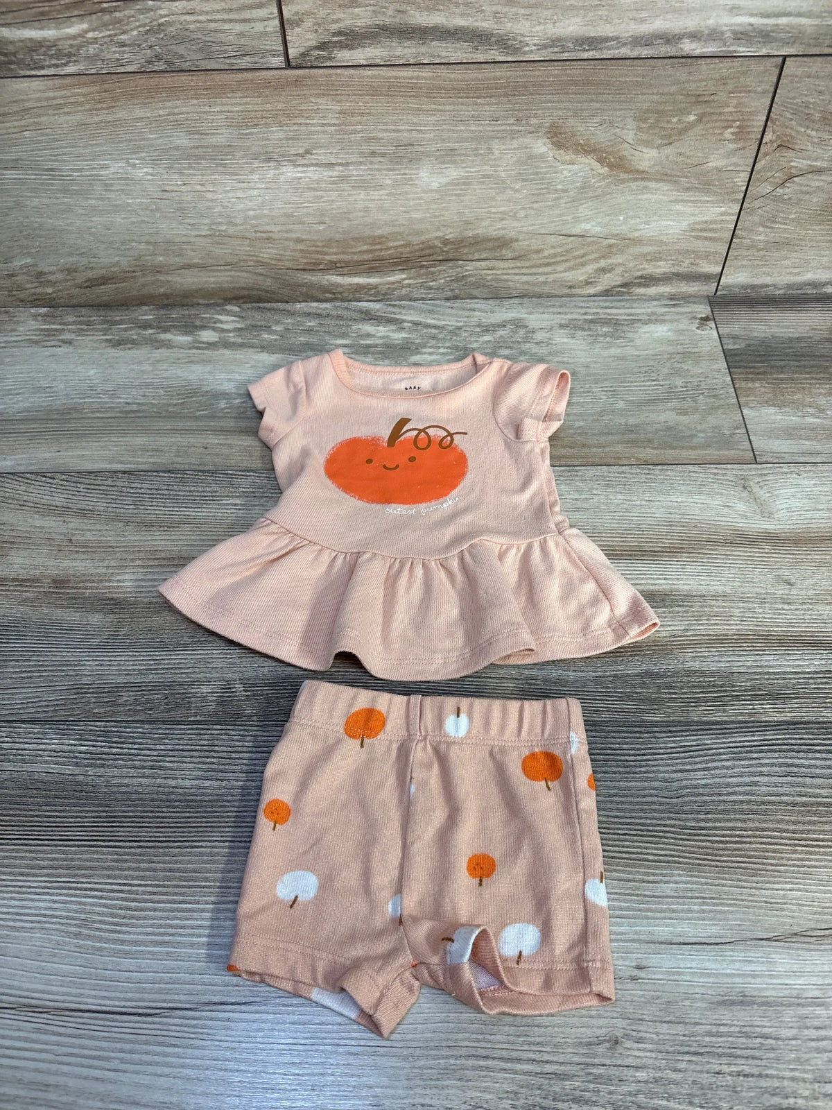 Flat lay of orange Cat and Jack pumpkin top and shorts set showcasing peplum detail and pumpkin print on wood-look floor