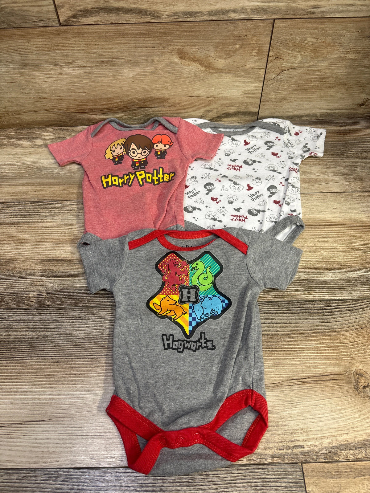 Front view of Harry Potter three-pack bodysuits in grey, red, and white with character and Hogwarts graphics on a wood surface