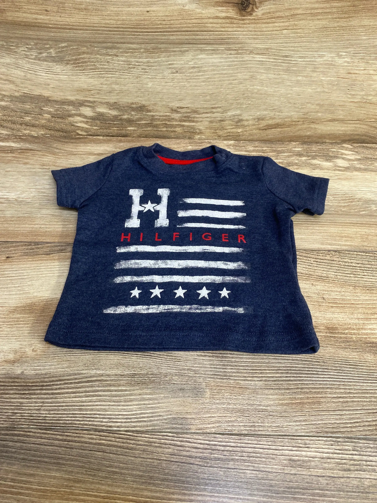 Front view of navy Tommy Hilfiger baby flag t-shirt showing white distressed stripes, stars, and red HILFIGER text on wood background