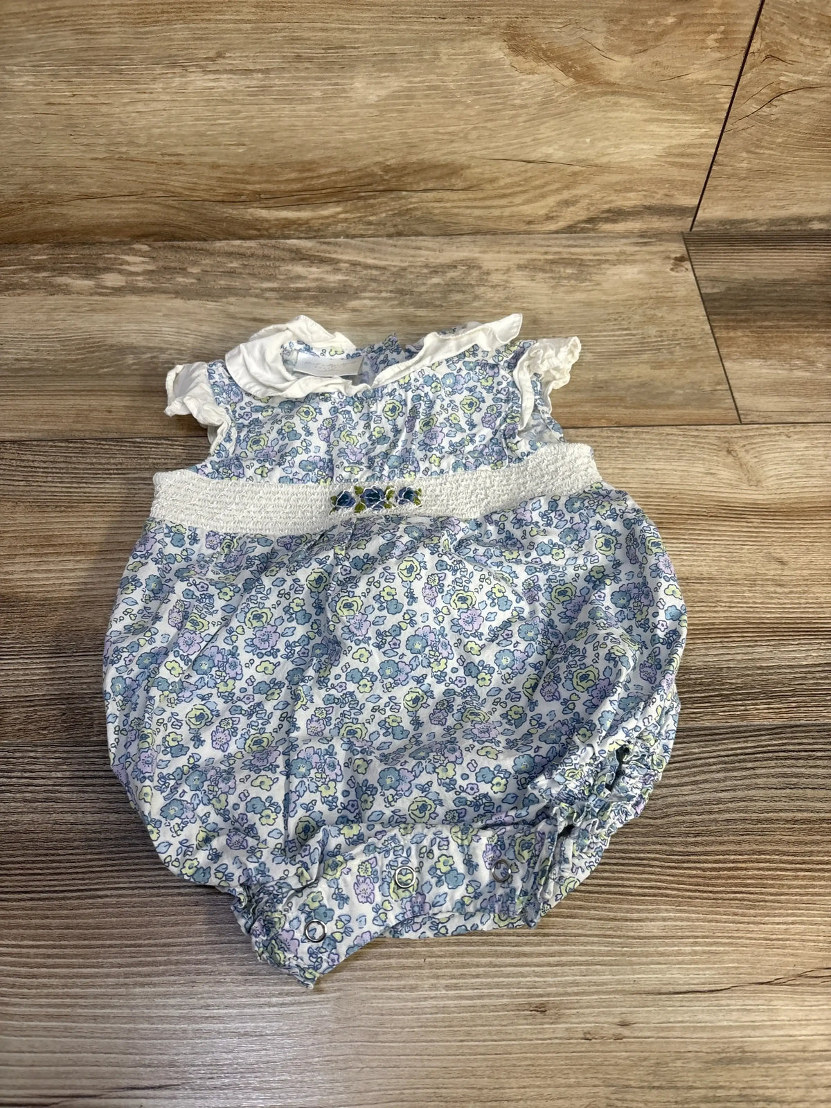 Top down view of white floral embroidered bubble romper showcasing smocked waist and ruffled sleeves on wood background